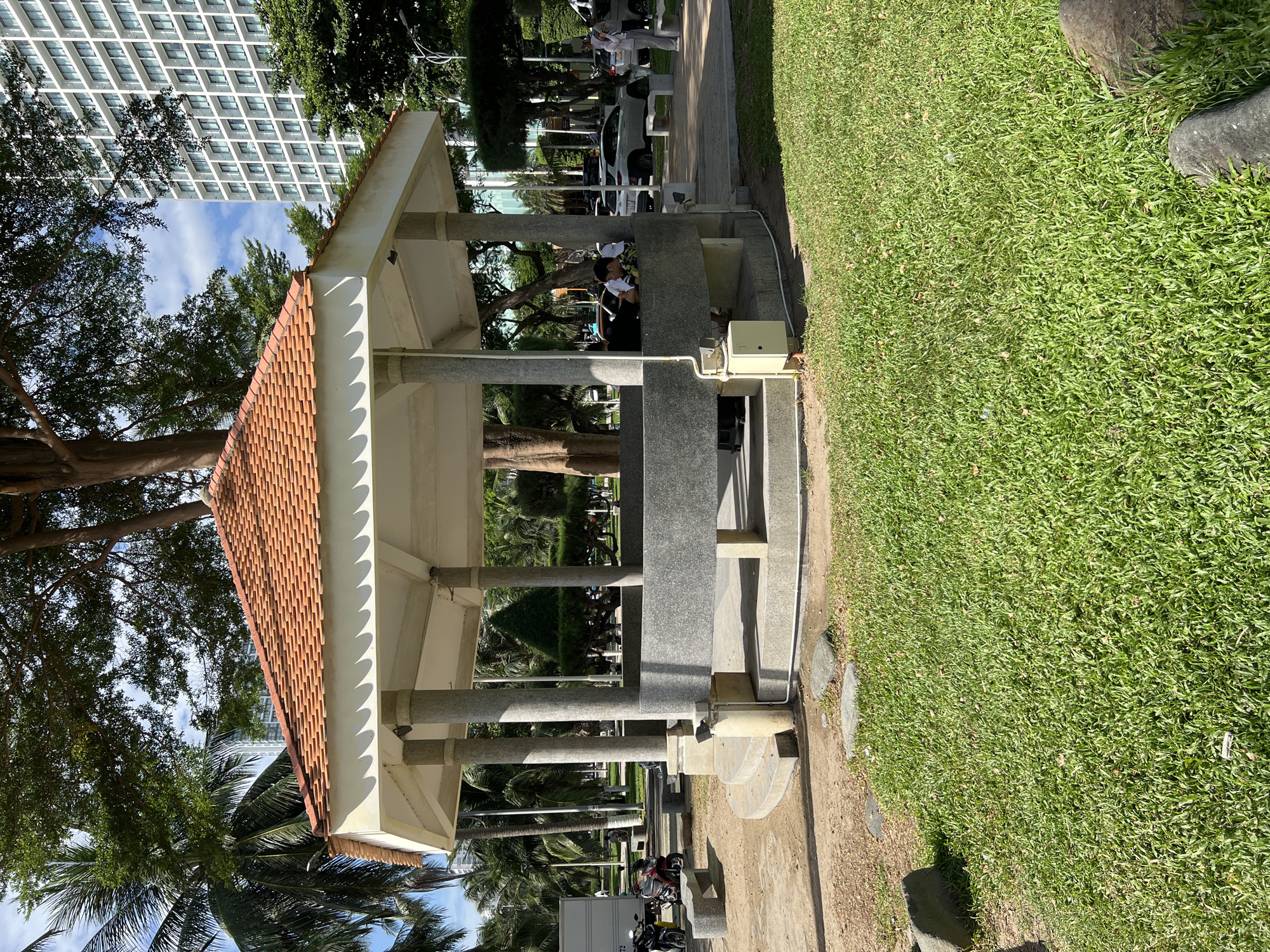 This image depicts an octagonal gazebo located in a park in Nha Trang, Vietnam. The gazebo features a terracotta tiled roof supported by stone pillars. Within the gazebo, two individuals are seated on a stone bench, one looking towards the camera. Outside the gazebo, to the left, two motorcycles are parked on a paved area. In the background, a modern high-rise building and lush palm trees are visible, suggesting an urban coastal environment. The ground is a mix of grass and paved pathways.