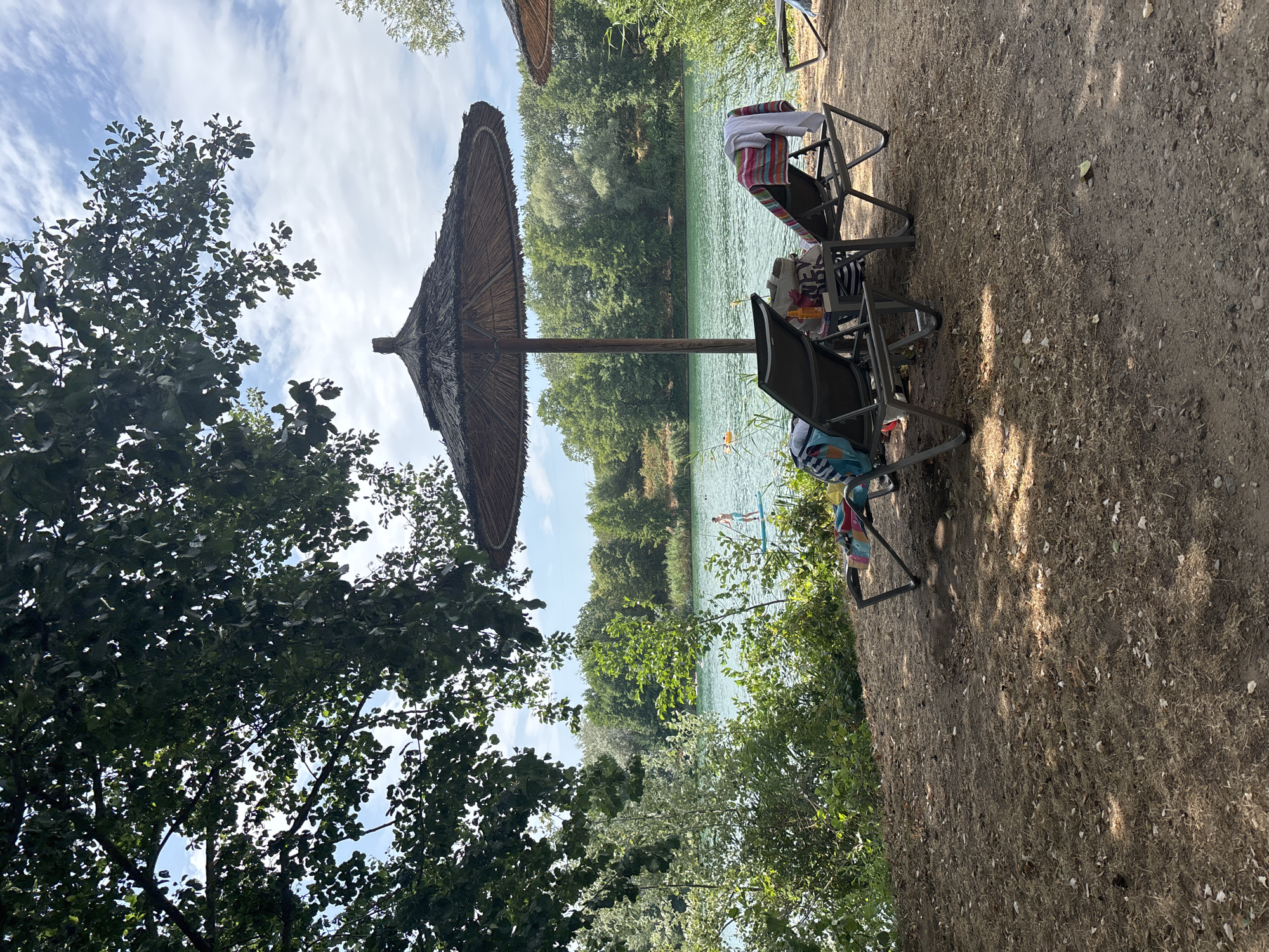 This is a photograph of a lake beach in Niedernberg, Germany. The scene depicts a serene lake with lush green trees lining the opposite shore. In the foreground, there are two empty lounge chairs and a large thatched umbrella providing shade. A towel is draped over one of the lounge chairs, and a bag and sunscreen sit on a small table between the chairs. In the distance, a person is visible on a paddleboard in the lake. The sky is partly cloudy.