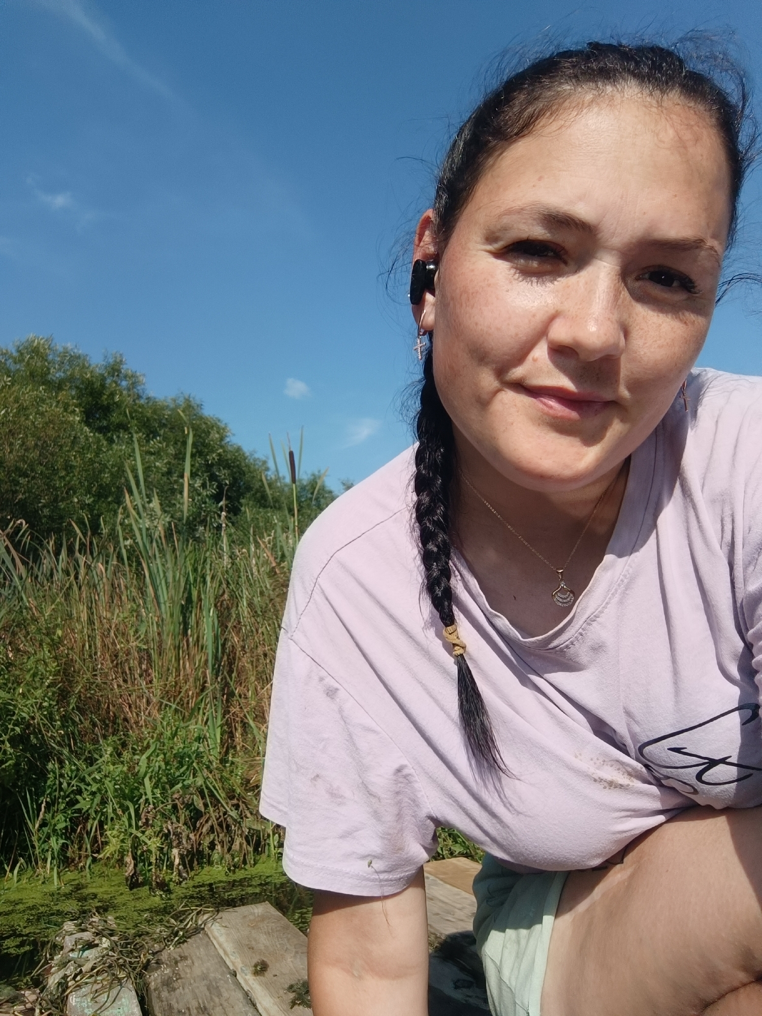 A woman is taking a selfie outdoors. She has dark hair in a braid and is wearing a light purple t-shirt, a necklace, and earrings. She is also wearing black earbuds. Behind her, there is lush green vegetation and a bright blue sky. The setting appears to be a natural environment, possibly a park or a rural area.