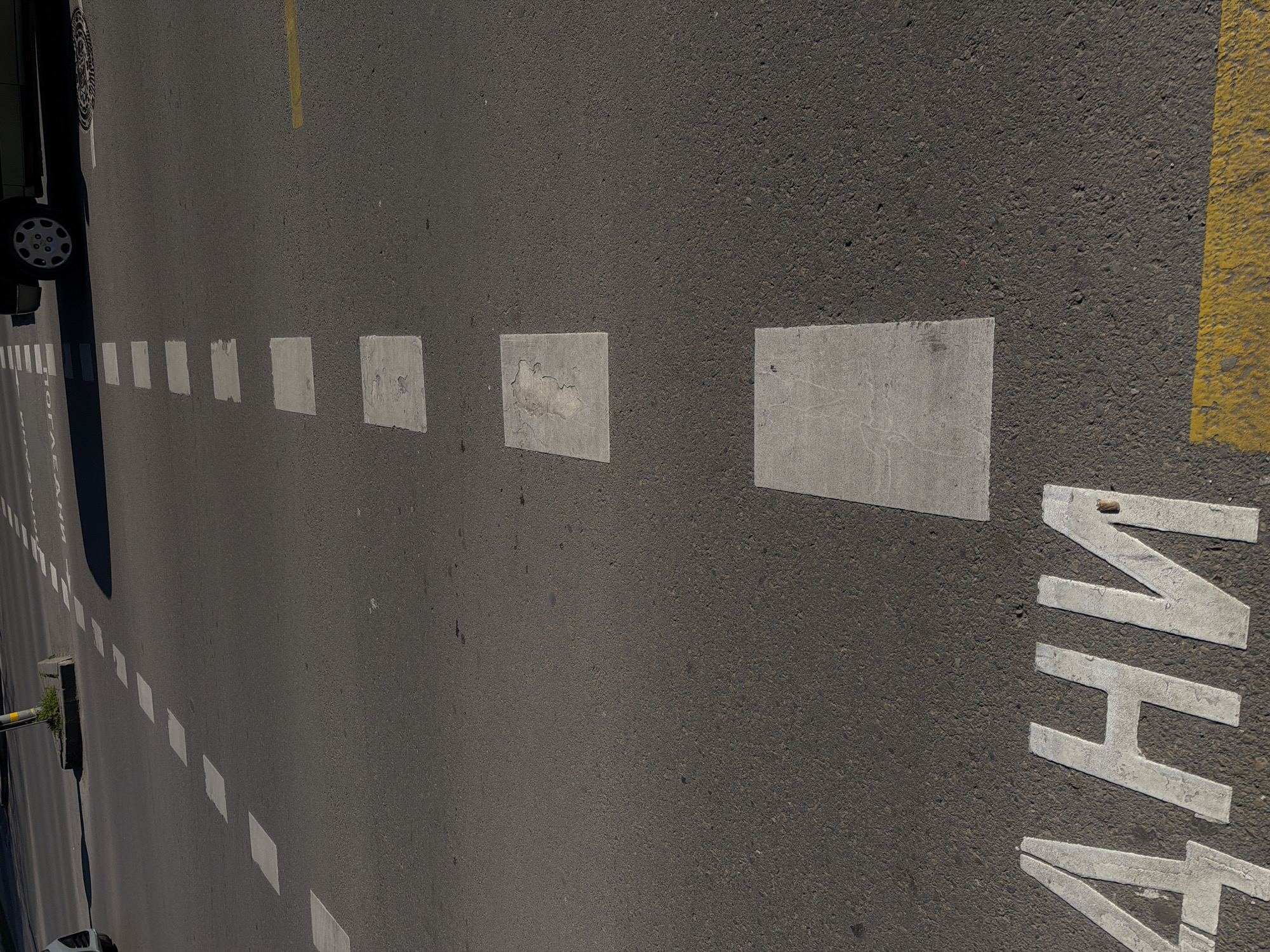 The image depicts an asphalt road surface in Varna, Bulgaria, marked with white and yellow traffic lines and road markings. The road is gray and shows signs of wear. A yellow line is visible on the right side of the image. To the left of it, there are several rectangular white road markings. On the left side, a car is parked along the road. White dashed lines are also visible on the left side. Road markings spelling out "BHM" are seen in the lower right portion of the image. The entire scene is bathed in sunlight, casting shadows.