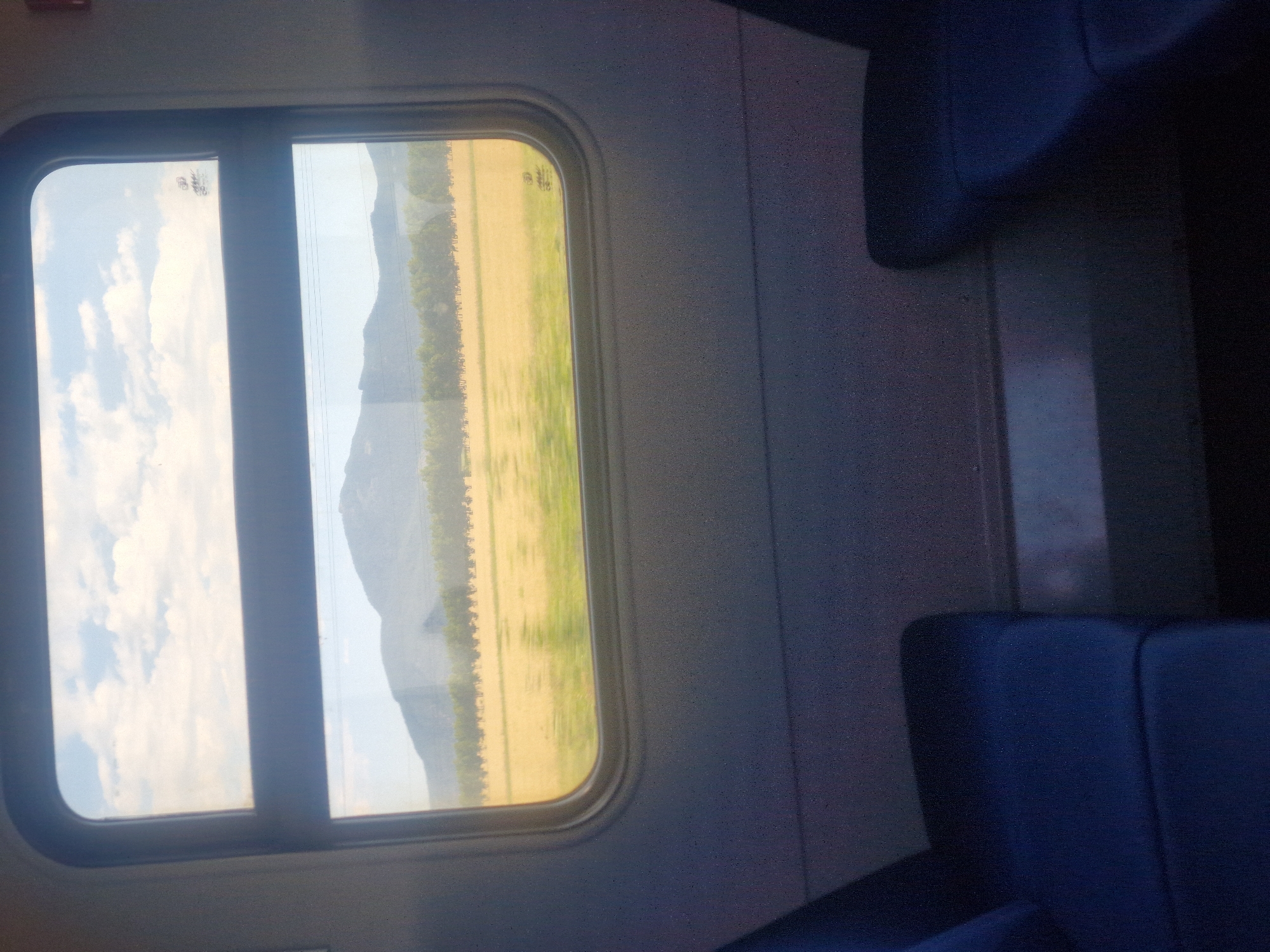 The image shows a view through a train window. Outside the window, a sunlit field of yellow and green crops stretches towards a line of trees and distant mountains under a cloudy sky. Inside the train, two dark blue seats are visible in the foreground, one on the left and one on the right, with the space between them suggesting a passage. The scene is likely from a train traveling through the rural landscape of Кенесары ауылдық округі, Kazakhstan.