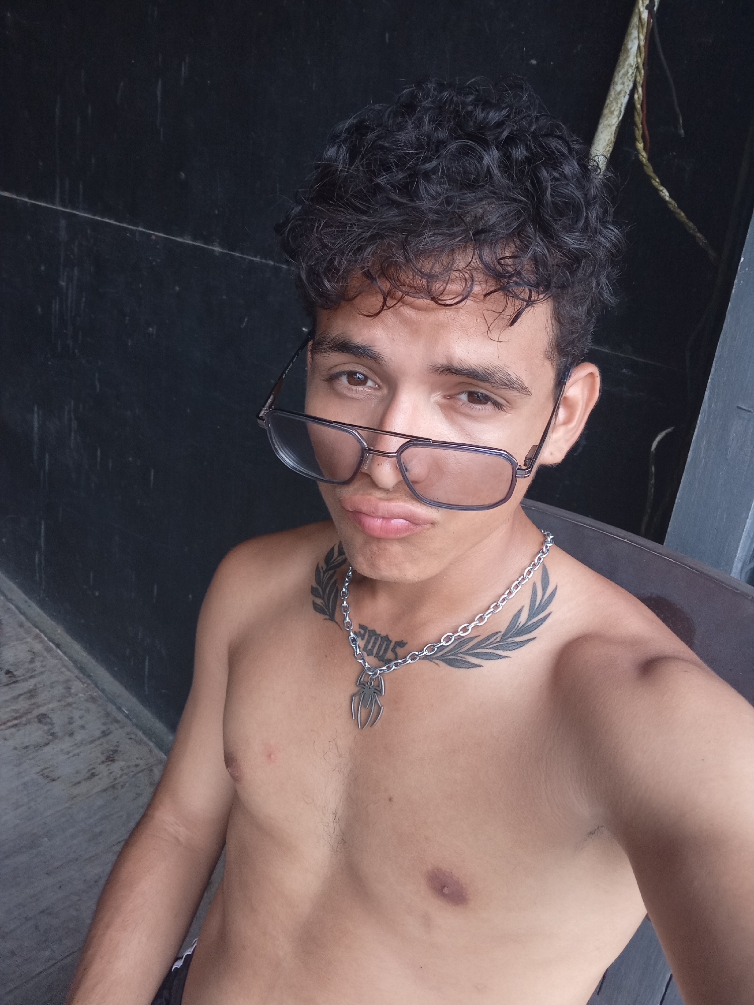 A young, shirtless individual with curly dark hair and brown eyes poses for a selfie. They are wearing glasses with rectangular frames, a silver chain necklace with a pendant, and have tattoos on their upper chest and neck. The background is a dark, possibly weathered wall, with a hint of vegetation or a rope visible in the upper right corner. The location is in Playa El Agua, Venezuela.