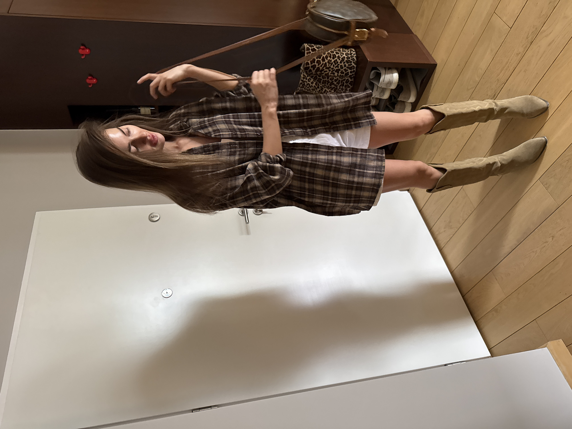 A woman is indoors, holding a brown leather handbag in front of her. She's wearing a plaid shirt over white shorts and suede cowboy boots. The setting appears to be a room inside an apartment, evident from the white door and wooden floor. The location of Unknown, Unknown cannot be confirmed.