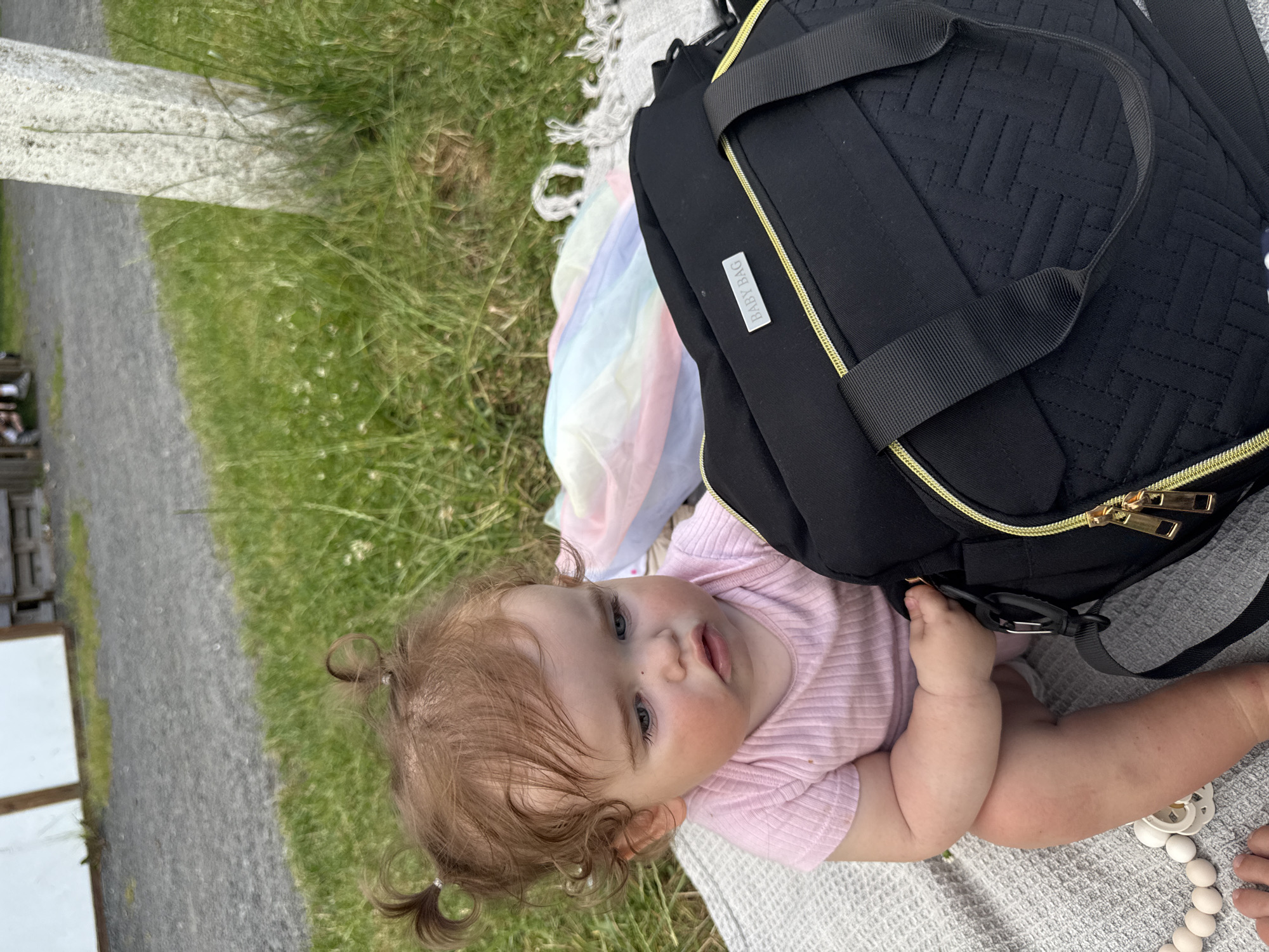 A baby with light brown curly hair styled into two pigtails sits on a grey blanket next to a black diaper bag. The baby is wearing a pink ribbed onesie and has blue eyes. The diaper bag has gold zippers and a silver "BABY BAG" tag. Behind the baby and bag, a colorful, folded umbrella is visible on the blanket. The scene is outdoors on a grassy area, with a gravel path in the background. A white pillar stands to the right of the frame. The location is not in Lumda, Germany.
