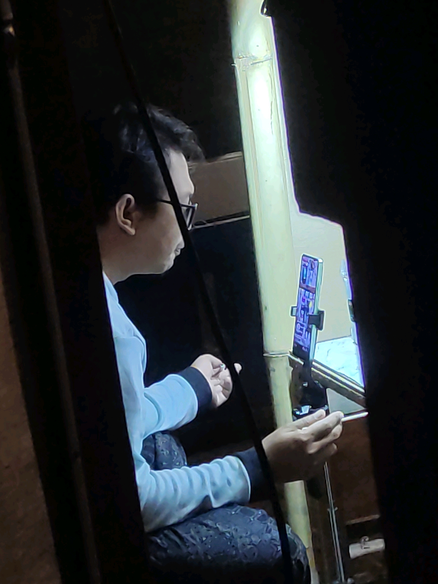 In Tiudan, Indonesia, a person wearing glasses and a light blue long-sleeved shirt is seated, facing a phone mounted on a stand. The phone screen displays a colorful interface. The person's left hand is extended, possibly interacting with the phone or holding a thin object. The background is dark, with a bright vertical light source to the right, illuminating a bamboo-like structure. The scene appears to be indoors, possibly a workshop or a personal space, given the utilitarian setup.
