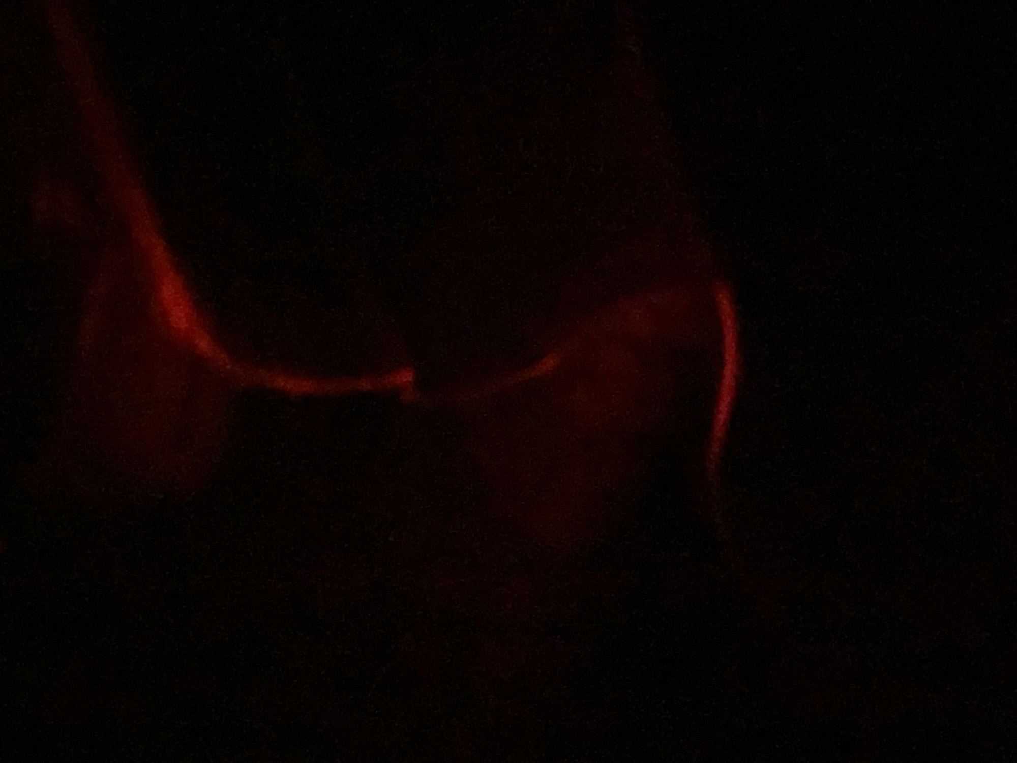 The image depicts a dimly lit, dark environment with a faint red glow emanating from the center and extending towards the right. The red glow appears to be the illumination source or a prominently featured object, with some amorphous shapes visible within or near the light. The background is predominantly black, suggesting a lack of ambient light. There is no identifiable urban landscape, structures, or features of Novokuznetsk, Russia. The scene is abstract and lacks discernible subjects, objects, actions, or interactions.