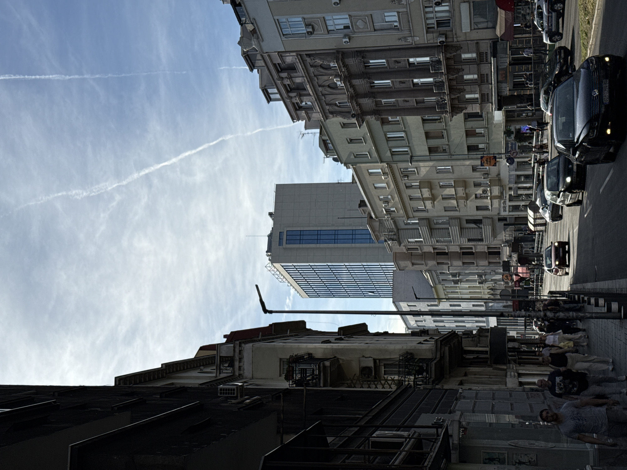 The image shows a street view in Belgrade, Serbia. Buildings line both sides of the street, with varying architectural styles and heights. A modern glass-faced building stands alongside older structures. Pedestrians are walking on the sidewalk, with several people in the foreground and others further down the street. Cars are parked along the right side of the street. The sky is visible above the buildings, with contrails from airplanes. The overall light suggests it is daytime.