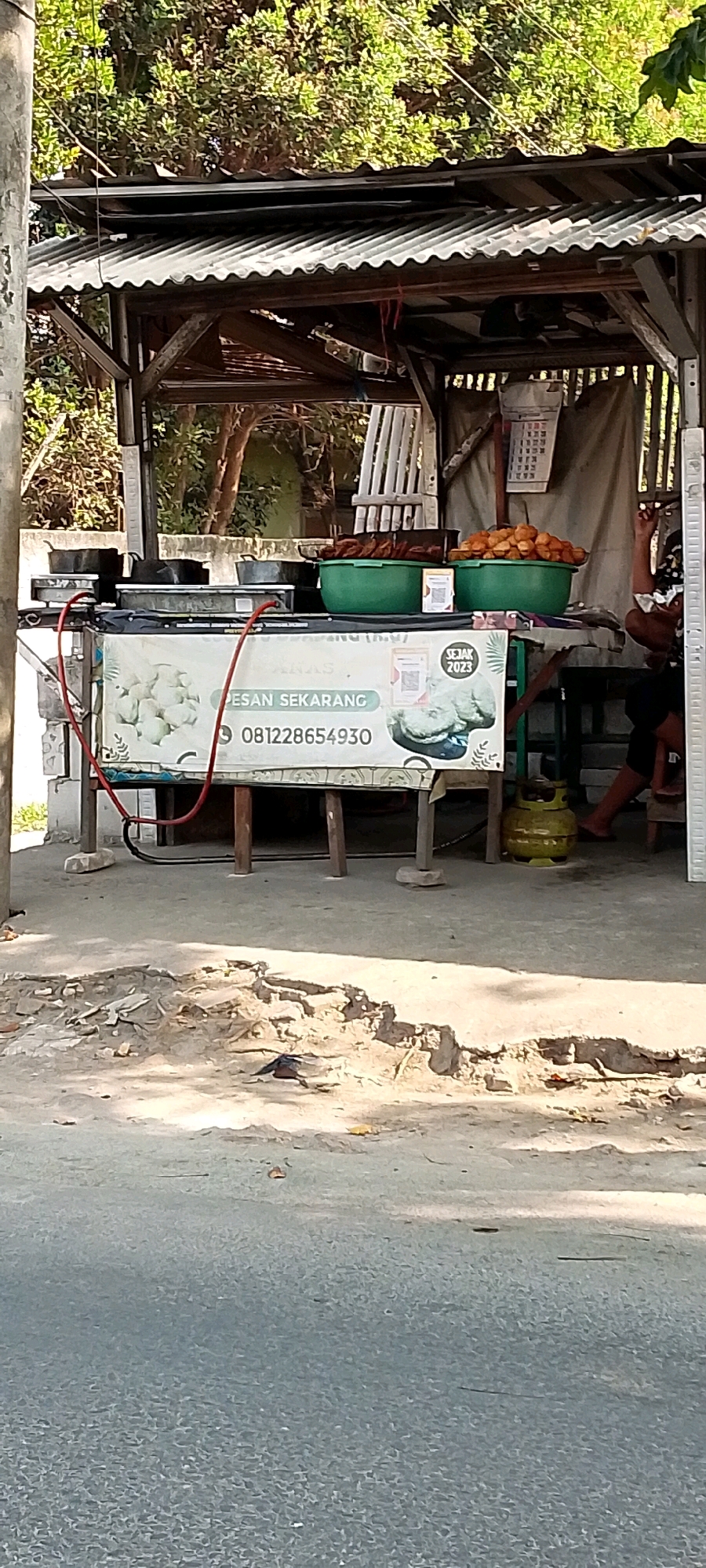 In Babelan Kota, Indonesia, a roadside food stall is pictured, constructed with a corrugated metal roof supported by wooden beams. The stall displays a banner with the text "PESAN SEKARANG" and a phone number "081228654930," along with imagery of fried food. In front of the stall, two green bowls are filled with fried items, and there are several pots and pans on a flat surface. A yellow gas cylinder is visible near the right side of the stall. The ground in front of the stall is paved, with some debris scattered on it. In the background, trees and foliage are present. A person's legs and arm are partially visible seated behind the stall.