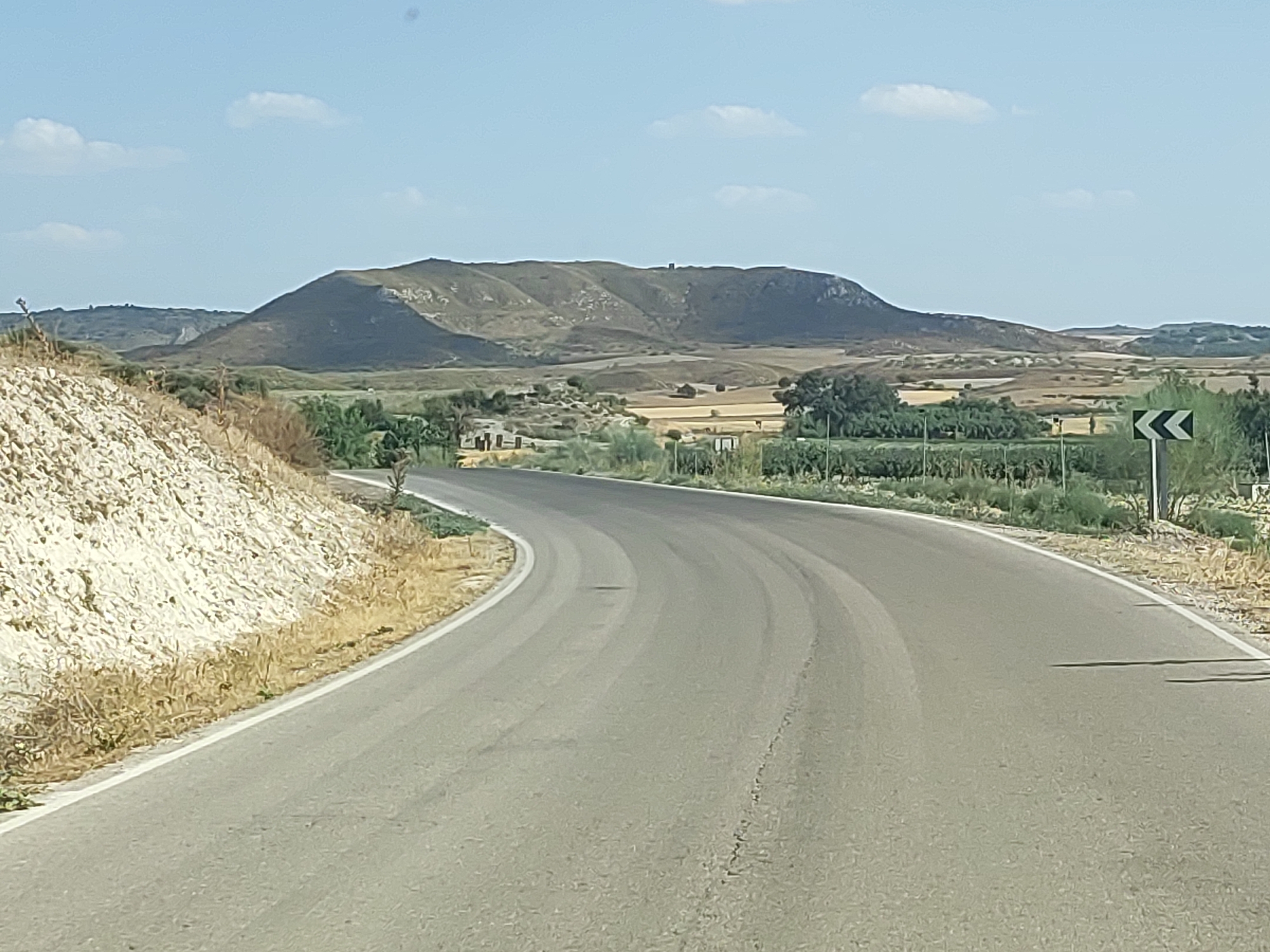 The image depicts a curved asphalt road with white lane markings leading through a rural landscape. On the left, a steep, light-colored, rocky embankment covered in dry grass rises from the roadside. The road appears to be in fair condition with some minor wear and tear. In the distance, rolling hills covered in dry vegetation stretch across the horizon under a pale blue sky with scattered white clouds. Several clusters of trees and low-lying vegetation are visible in the mid-ground, along with patches of tilled or harvested fields. A single road sign with black chevron markings on a white background is positioned on the right side of the road. There are no people or vehicles clearly visible in the image. This scene is characteristic of the arid, hilly terrain found in and around Illana, Spain.