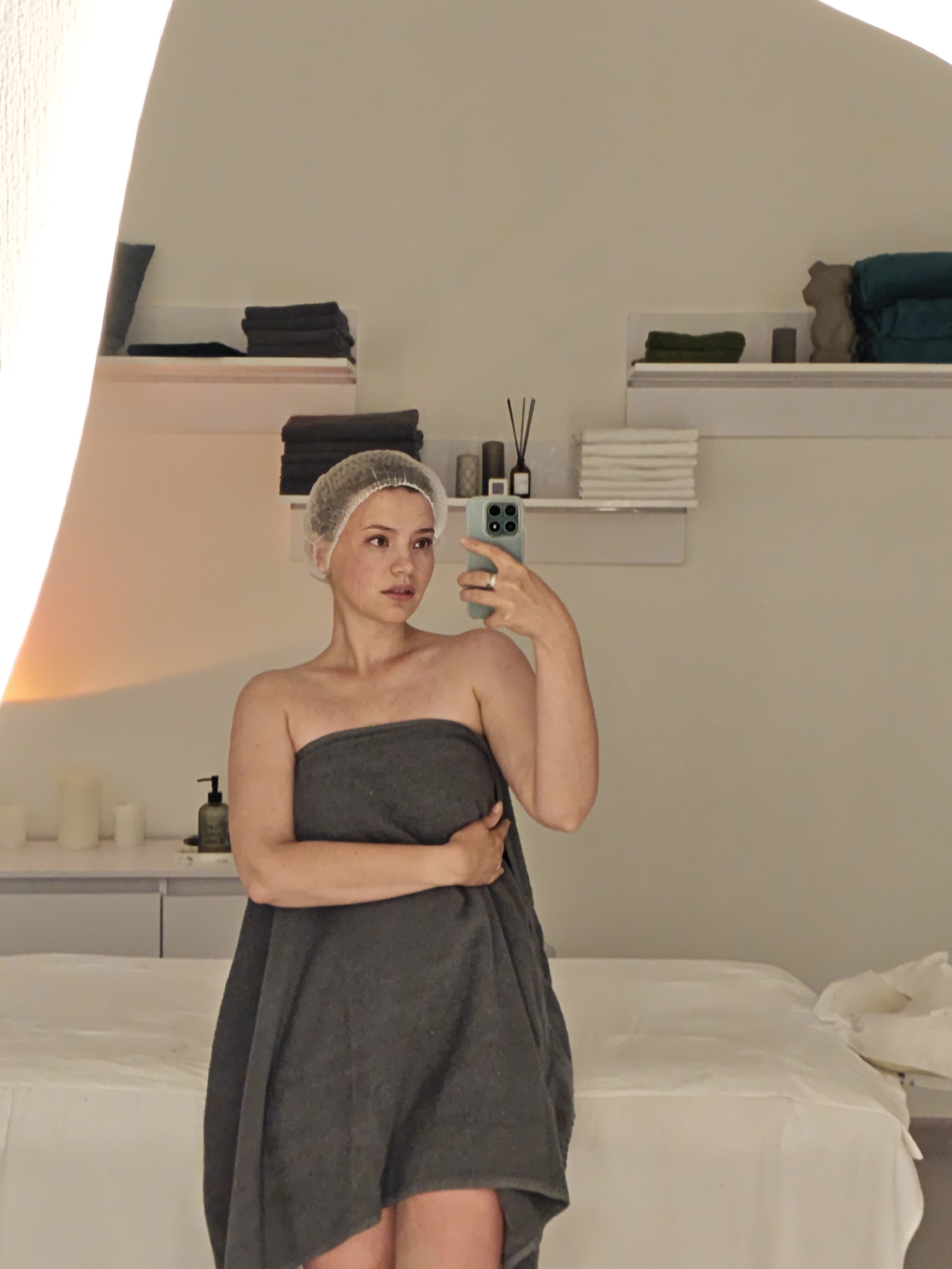 A young woman with a cap on her head and wrapped in a dark gray towel is taking a selfie in a spa room. She is holding a light blue smartphone in her right hand and her left arm is bent, resting on her chest. A massage table is visible behind her, covered with a white sheet. Shelves on the wall hold folded towels and decorative items. The room is lit by artificial light, casting a warm glow on the scene. The location is identified as a spa in Vinnytsia, Ukraine.