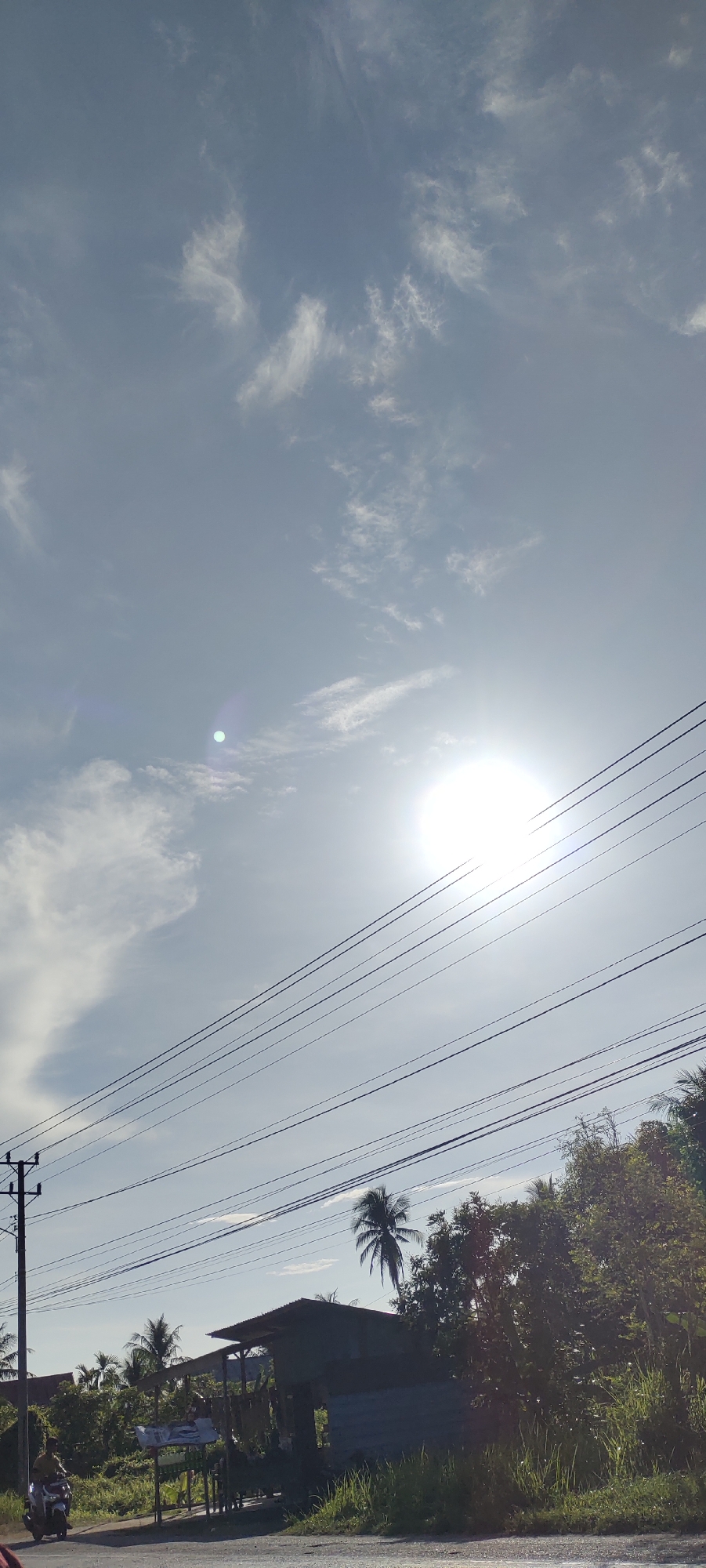 In Matangpanyang, Indonesia, a bright sun shines in a partly cloudy sky. Multiple utility wires are strung across the frame, supported by a utility pole on the left. Below the wires, a small, dark-colored building with a corrugated metal roof is visible, set back from a road. A person on a white motorcycle is approaching the building on the left side of the road. Lush green vegetation, including trees and grass, surrounds the building and the roadside. The scene appears to be a rural or semi-rural area.