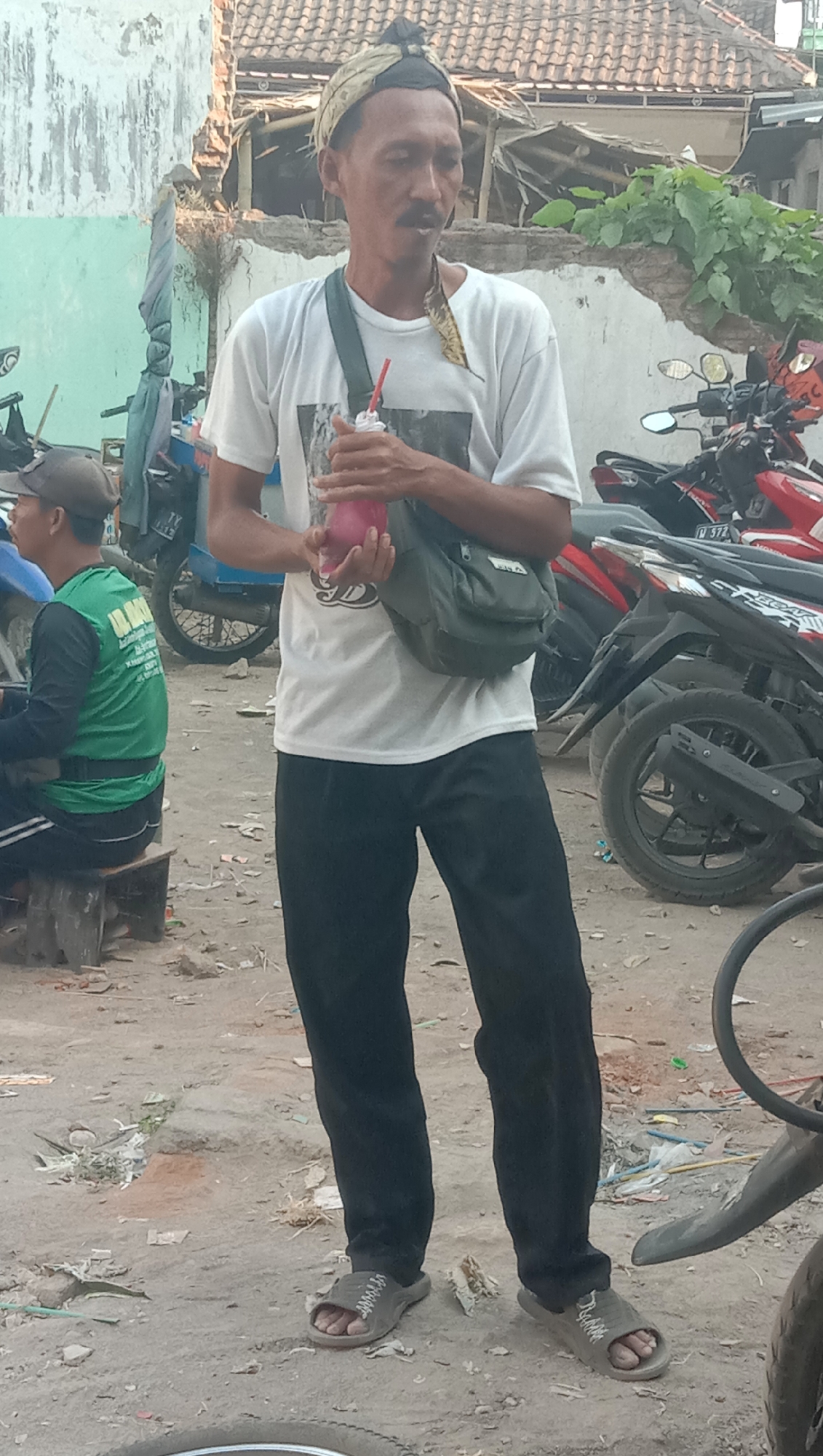 The image depicts a man standing in what appears to be an outdoor marketplace or a street in Sidogiri, Indonesia. He is wearing a white t-shirt with a graphic print, black pants, and gray slip-on sandals. A green shoulder bag is slung across his chest, and he is holding a pink beverage in a plastic cup with a red straw. The man has a mustache and is wearing a traditional Indonesian headband.
In the background, there are motorcycles parked. To the left, another person is seated on a wooden stool, wearing a green long-sleeved shirt and a gray cap, facing away from the camera. The environment is dusty and appears to be a commercial area, with a building with a tiled roof and some debris on the ground. The overall scene suggests a casual, everyday setting within a populated area.