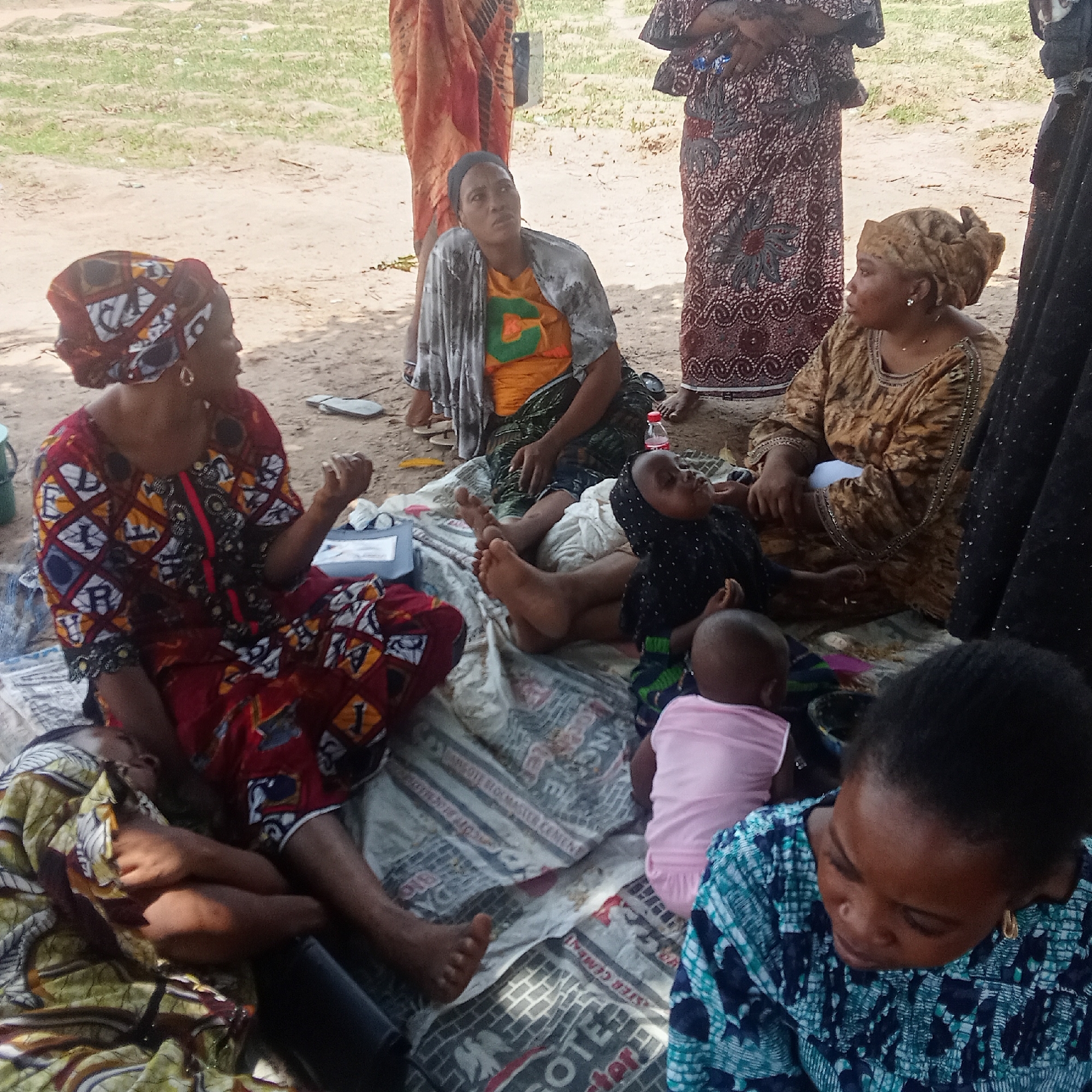 A group of women and children are gathered outdoors in Doma, Nigeria. They are sitting on a patterned blanket spread on the ground. The women are wearing colorful traditional clothing. One woman in a red patterned dress is talking while gesturing with her hand. Another woman in a yellow dress is looking at her. A third woman in a bright orange shirt looks upward. Several babies and children are lying or sitting on the blanket. The ground is sandy.