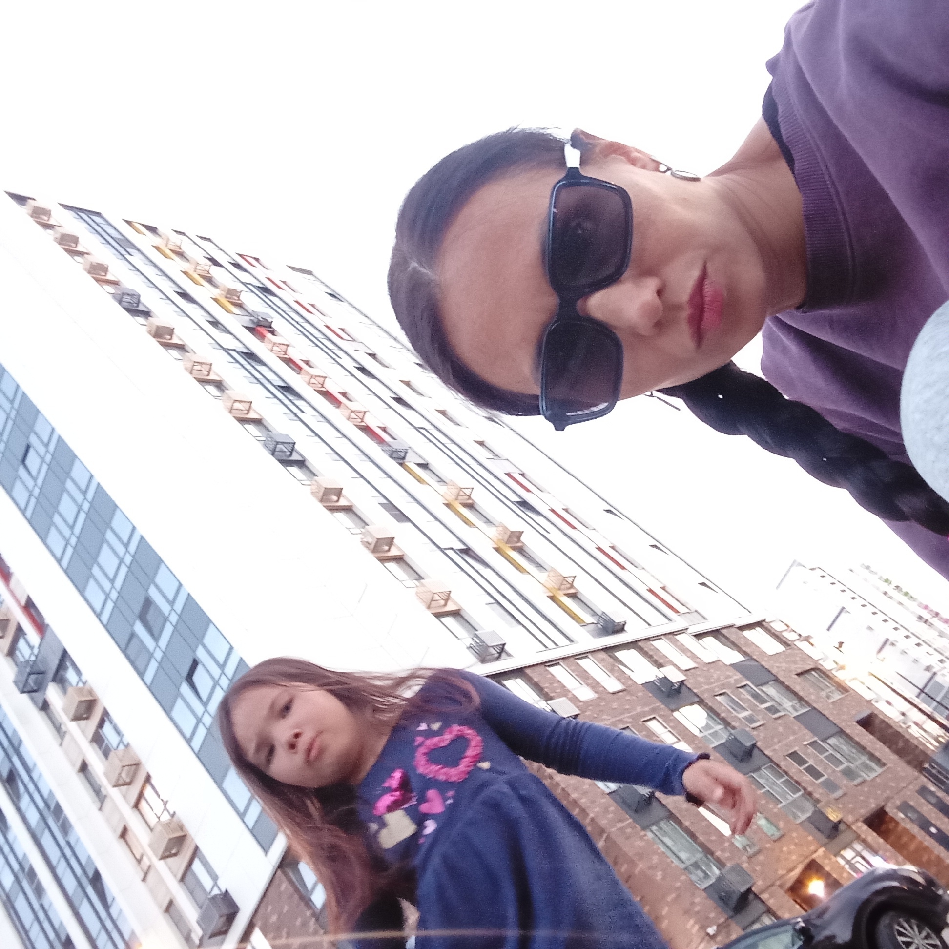 A low-angle, upward-tilted photograph captures an adult woman and a young girl positioned against a backdrop of modern high-rise residential buildings under an overexposed white sky. The scene is located in Izhevsk, Russia.
In the upper right foreground, an adult woman is visible, looking downwards towards the camera. She has dark hair, styled with a center part and a visible dark braid extending over her right shoulder. She wears large, rectangular dark sunglasses, obscuring her eyes, and a purple crew-neck sweatshirt with a subtly textured, possibly velvet, fabric. Her lower lip shows red-pink lipstick.
To the lower left and slightly behind the woman, a young girl stands, looking generally towards the camera but not directly. She has long, light brown hair and is dressed in a dark blue long-sleeved dress adorned with multiple pink and light-colored heart motifs across the chest. Her right arm is slightly extended forward.
The background features at least two distinct multi-story apartment buildings. The prominent building on the left half of the frame has a predominantly white facade with numerous rectangular windows, some appearing reflective blue. It features a repeating pattern of small, square or rectangular, light-brown, and dark-grey protrusions, likely housing external air conditioning units or decorative elements, arranged vertically along its structure. To the right, another building with a reddish-brown brick facade is partially visible, also featuring multiple windows and some yellow/orange external fixtures. A dark-colored vehicle, possibly an SUV, is partially visible in the bottom right corner, showing a tire and part of its body, suggesting a street or parking area at ground level.