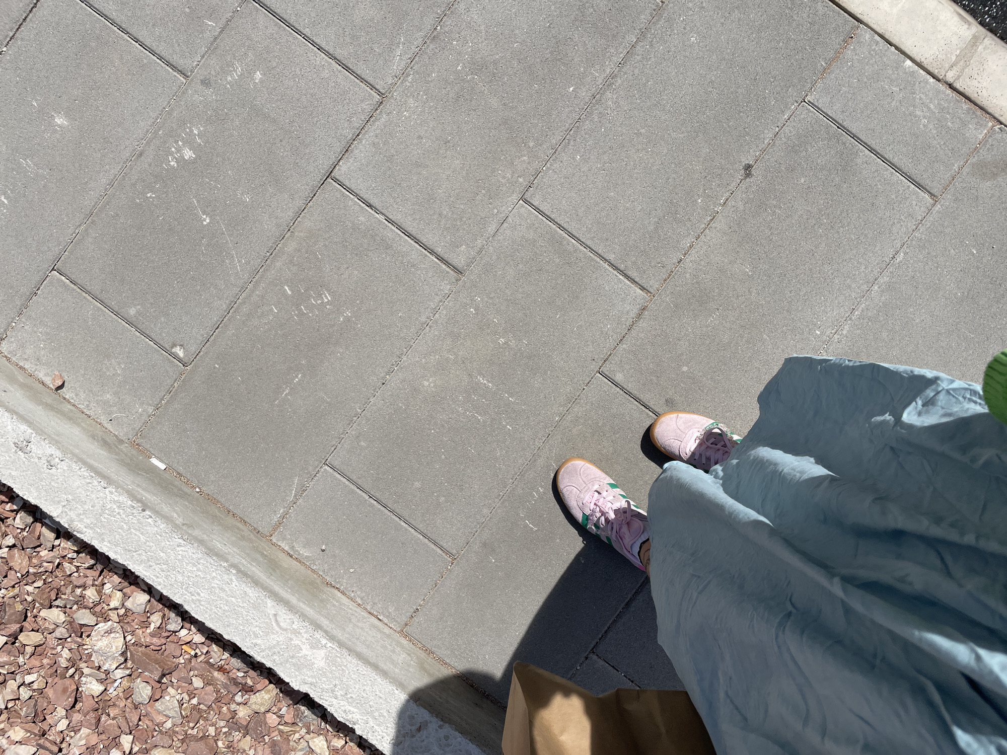 The image shows a first-person perspective of a person standing on a gray paved sidewalk, potentially in Rafailovići, Montenegro, though this location cannot be confirmed from the visual data alone. The paving consists of rectangular stones. The person is wearing light pink sneakers with green stripes. A long, light blue dress or skirt covers most of the lower right portion of the image. Part of a brown paper bag is also visible. To the left of the sidewalk is a stone curb followed by a bed of reddish-brown rocks or gravel. The lighting suggests it is daytime.