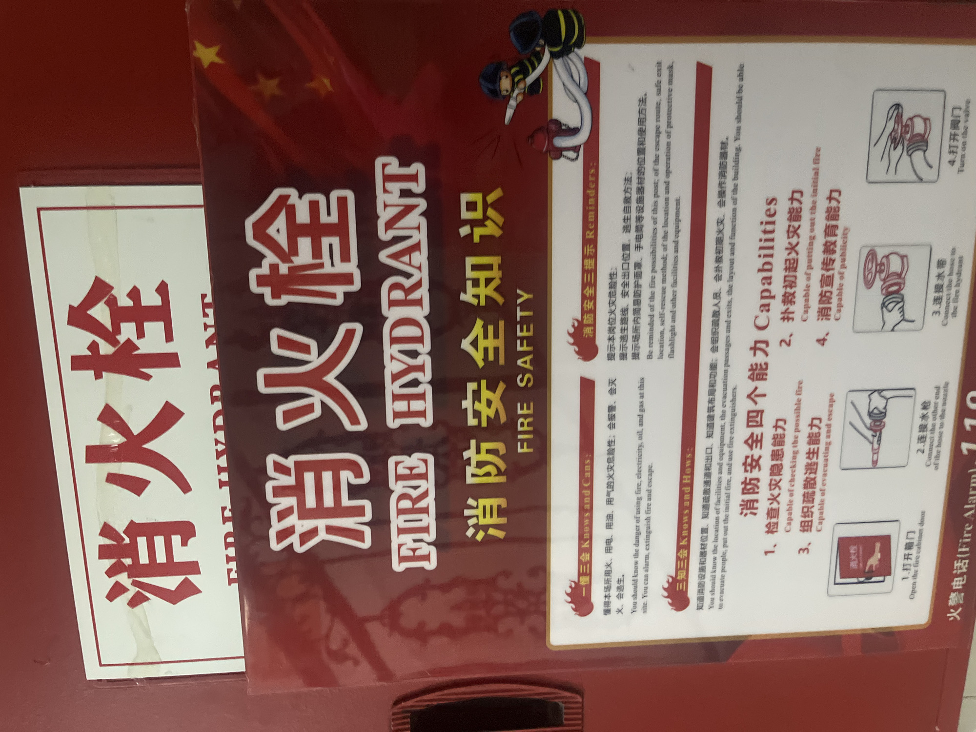 The image displays a red fire hydrant access panel or cabinet door, prominently featuring fire safety information. At the top, a smaller sign with a white background bears the Chinese characters "消火栓" (Xiāohuǒshuān) and the English text "FIRE HYDRANT". Directly below, on the red surface, a larger, slightly embossed graphic repeats "消火栓" and "FIRE HYDRANT", followed by "消防安全知识" (Xiāofáng ānquán zhīshì) and "FIRE SAFETY".
The red background is adorned with faint golden stars in the upper right quadrant, reminiscent of the Chinese national flag. To the right of the "FIRE SAFETY" text, two cartoon firefighters are depicted: one aims a hose connected to a red fire hydrant, while the other stands nearby, ready.
Below this, a white laminated sheet contains detailed fire safety instructions in both Chinese and English, divided into several sections:
* **"一懂三会 Knows and Cans:"** details the danger of fire from various sources (fire, electricity, oil, gas) and the abilities to alarm, extinguish fire, and escape.
* **"消防安全三提示 Reminders:"** lists reminders about fire possibilities, escape routes, safe exit locations, self-rescue methods, and the use of protective masks, flashlights, and other equipment.
* **"三知三会 Knows and Hows:"** outlines knowledge requirements regarding the location of facilities, equipment, evacuation passages, exits, building layout, and the ability to evacuate people, extinguish initial fires, and use extinguishers.
* **"消防安全四个能力 Capabilities:"** describes four key capabilities: checking for possible fire hazards, putting out initial fires, organizing evacuation and escape, and fire safety publicity/education.
Four small diagrams illustrate the steps for using a fire hydrant:
1. Opening the fire cabinet door.
2. Connecting the hose to the nozzle.
3. Connecting the hose to the fire hydrant valve.
4. Turning on the valve.
At the very bottom of the red surface, partially visible, is the text "火警电话(Fire Alarm): 119". The overall scene suggests a fire safety station or public information display within a building in Yuzhong District, China.