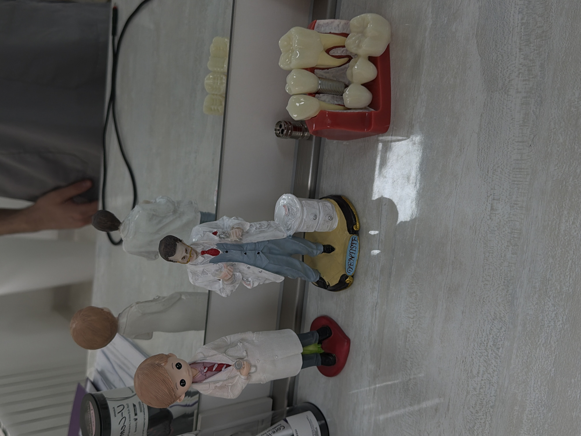 The scene is an interior setting, likely a dental office or clinic in Tbilisi, Georgia. The main subjects are figurines of dentists, placed on a light-colored countertop with a wood grain pattern.
One figurine stands on a red heart-shaped base, wearing a white coat, a red-striped tie, and holding a stethoscope. Another figurine is on a gold and black base labeled "DENTISTE". This figure also wears a white coat, with a blue vest underneath.
A dental model with several teeth, including implants, sits in a red tray. The model and figurines are positioned on a reflective surface, possibly a mirror or shiny countertop, which shows reflections of the objects, and a person’s arm and shoulder. There are also dental materials like creams. A thin black wire is visible near the mirror.