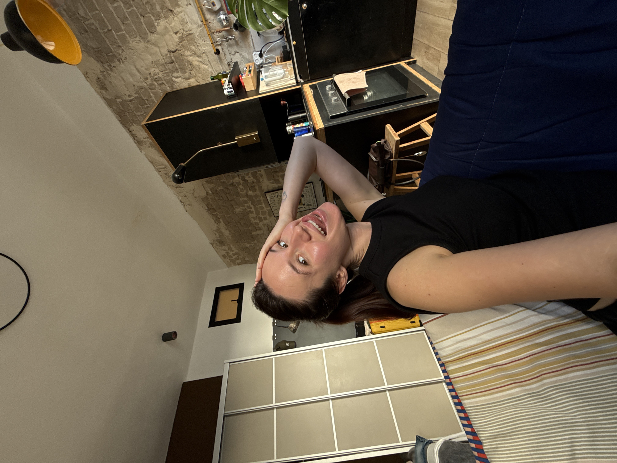 The image depicts a woman in a black t-shirt lying on a striped bed. She has her hand on her forehead and is smiling. The room has a white wall on the left with a black circular object hanging on it, a black framed picture and a small round object also on the wall. A closet with sliding doors is visible. There's also a brick wall ceiling. In the upper portion of the image are black cabinets and various items on top. The woman appears to be taking a selfie. Given the style of the interior, it's likely that the location is Tel-Aviv, Israel, a city known for its unique architecture and design elements.