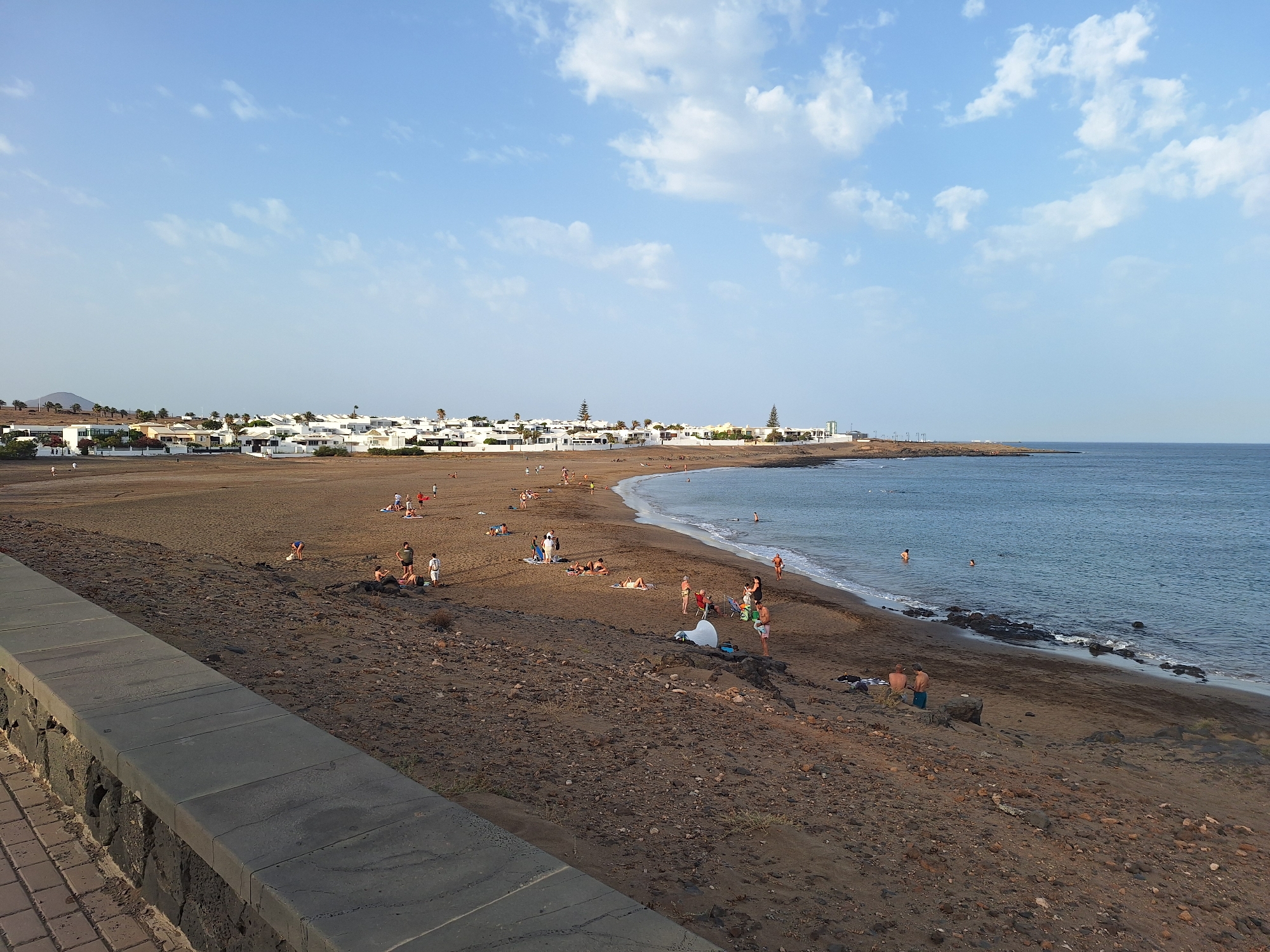The image captures a wide view of a beach in Arrecife, Spain. The foreground features a dark, textured stone wall and a brick-paved walkway. Beyond the wall lies a sandy beach, appearing dark brown or black, with scattered rocks and vegetation. The beach is populated by several individuals, some sunbathing on towels, others walking or standing. A few people are in the shallow water near the shore.
The sea, a calm blue, extends to the horizon under a pale blue sky with scattered white clouds. Along the coastline, white buildings with flat roofs form a townscape, interspersed with palm trees and occasional taller structures, possibly wind turbines or communication towers. The overall scene suggests a peaceful day at a coastal resort.