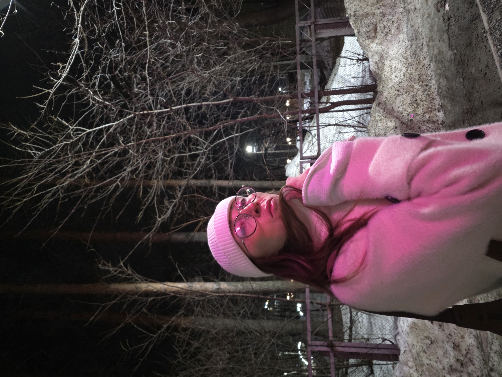 The image depicts a nighttime outdoor scene in the city of Chaikovsky, Russia, characterized by artificial illumination and winter conditions. In the foreground, a female individual is positioned slightly left of center, her body angled to her right, and her gaze directed upwards and to her right. She wears a white ribbed beanie, round-rimmed eyeglasses, and a textured, light-colored (appearing vibrantly pink due to reflective lighting) turtleneck or high-collared coat with visible dark buttons. Her left hand is raised towards her chin or neck, possibly adjusting her garment. The subject's face, beanie, and coat are strongly illuminated with a magenta-pink light, reflecting pink in her eyeglasses. Her hair, visible beneath the beanie, appears brown or reddish-brown.
The background features a densely treed area with numerous bare, dark-trunked trees and intricate networks of thin, leafless branches. Patches of dirty, melting snow cover the ground, interspersed with dark, exposed earth. A low, dark-colored fence or railing extends horizontally behind the subject. Multiple distant streetlights emit bright, white illumination, contrasting with the pink light on the subject. A wider, possibly wire-mesh fence is discernible further back in the treeline. The scene suggests a park or semi-urban forested area during a cold period, possibly late winter or early spring.