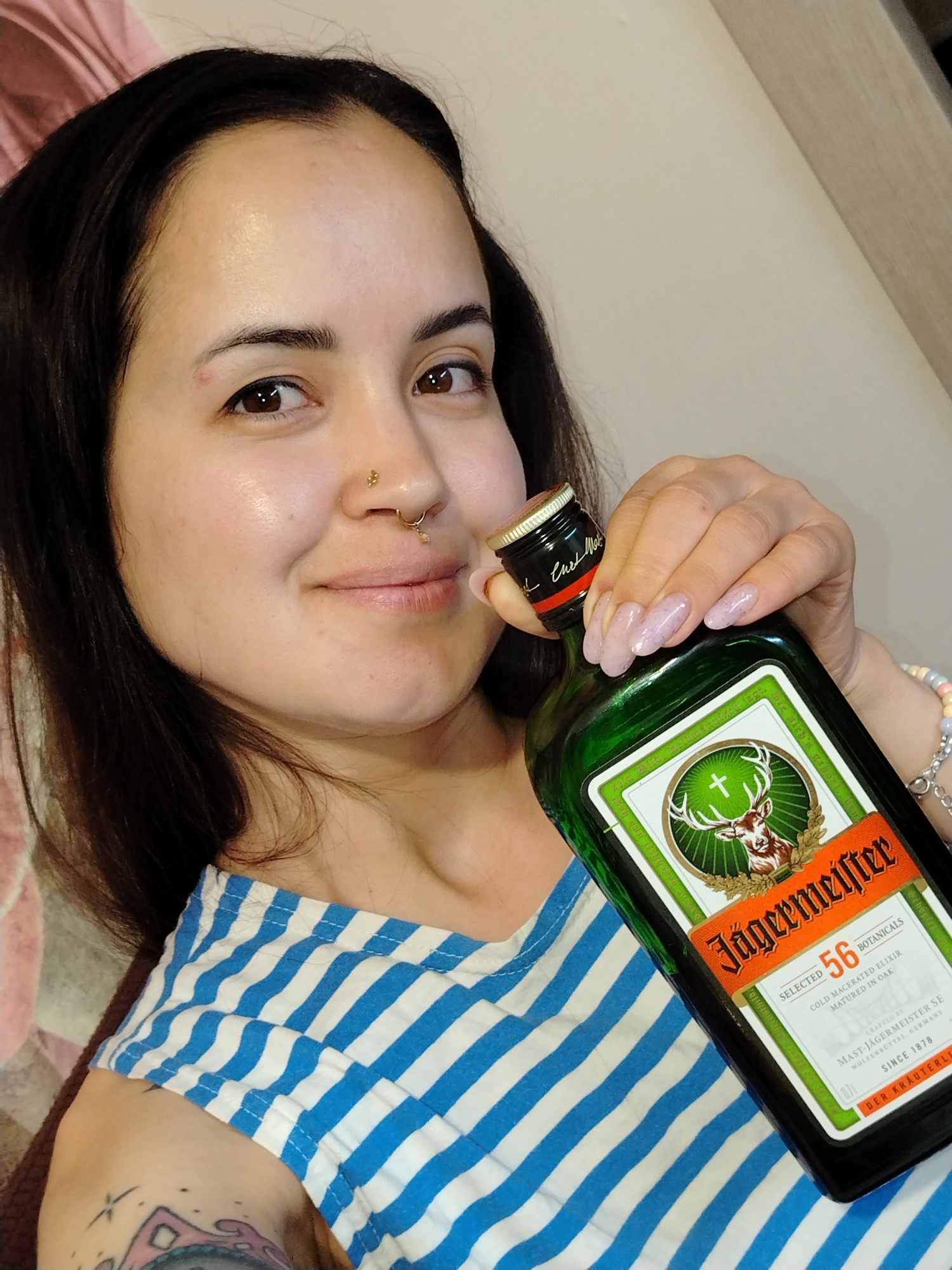 A woman is holding a bottle of Jägermeister. The woman has dark hair and is wearing a blue and white striped shirt. She has a nose piercing and a tattoo on her arm. The Jägermeister bottle is green and has a label with a deer on it. The label also states "SELECTED 56 BOTANICALS" and "MAST-JAGERMEISTER SE WOLFENSCTTEL, GERMANY". The scene is indoors. There is no indication of the location being in Noviy, Russia.