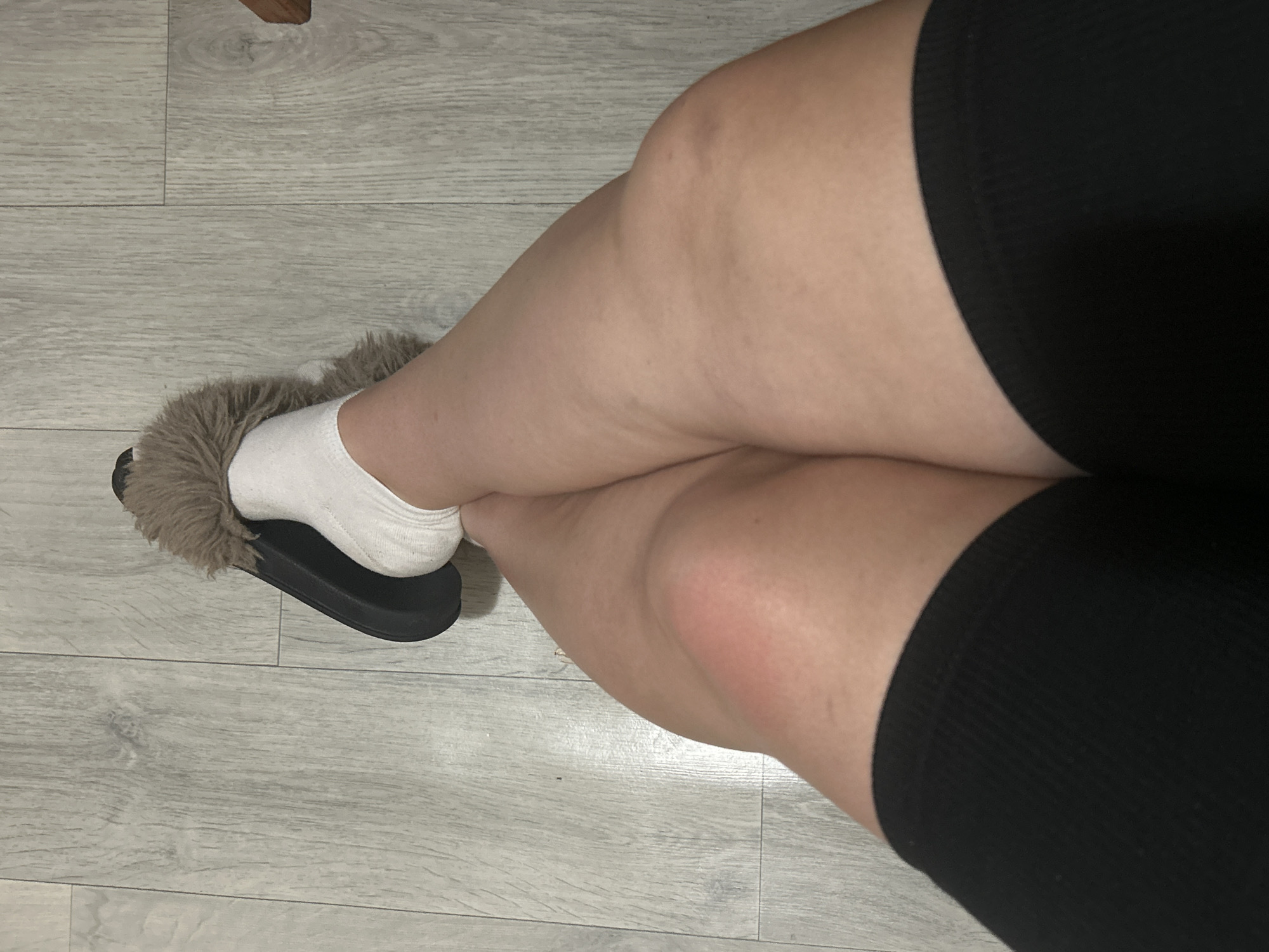 A person is sitting on a floor with their legs crossed, wearing black shorts, white socks, and fuzzy slippers. The floor is made of light-colored laminate wood planks. The image is taken from a high angle, looking down at the person's legs and feet. There is no information in the image to indicate its location in Basarabeasca, Moldova.
