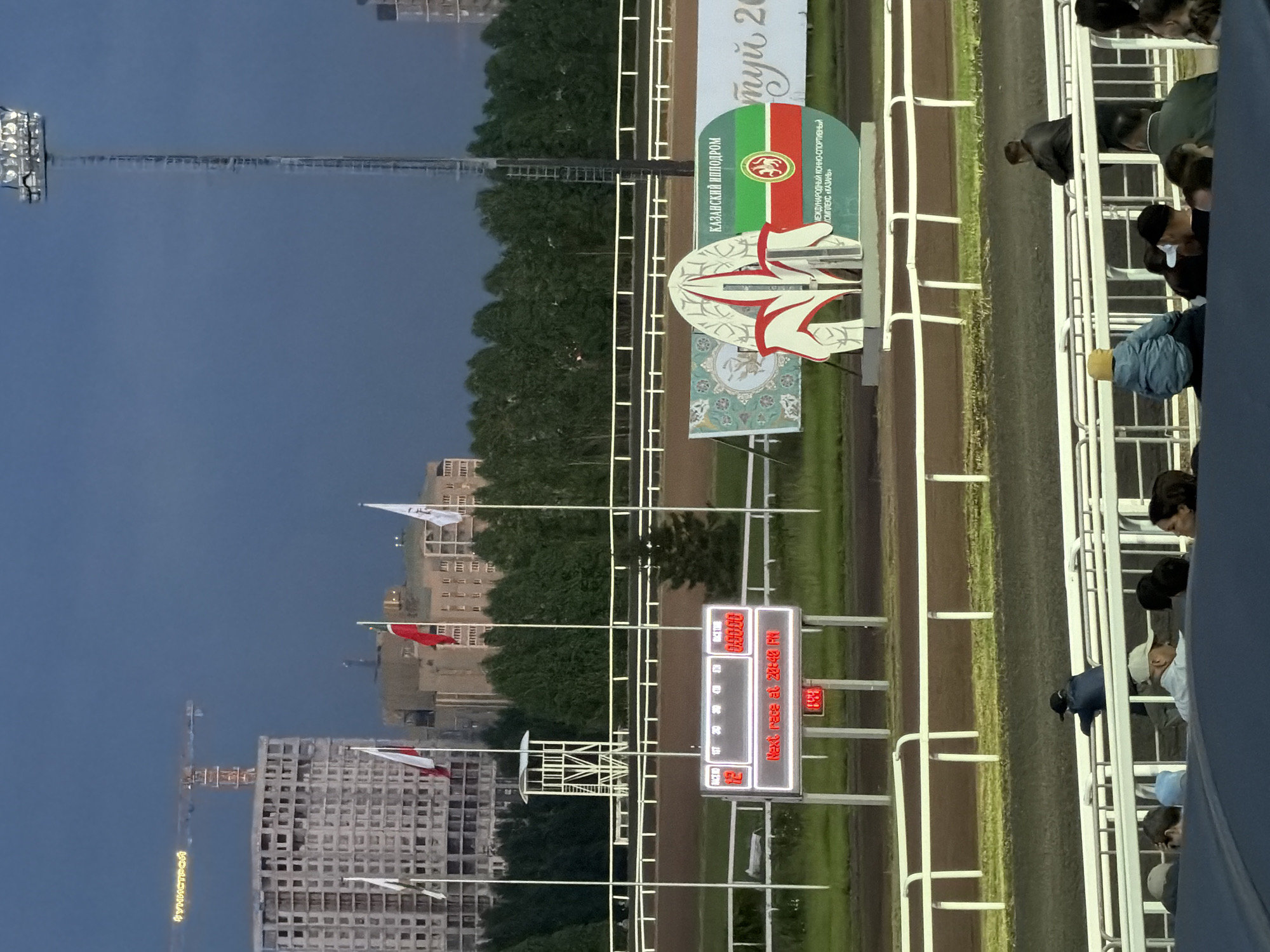 The image depicts an outdoor horse racing track at dusk. In the foreground, several people are seated in stands, some looking towards the track. A white fence separates the stands from the track, which is a dark dirt surface. In the mid-ground, a large electronic scoreboard displays race information, including "RACE NO 12" and "NEXT RACE AT 20:48 PM". To the right of the scoreboard, a large circular sign features the flag of Tatarstan, along with text that appears to be in Russian. Behind the track, a line of trees forms the background, with buildings and construction sites visible in the distance. A tall light tower with multiple spotlights stands to the right of the frame. The sky is a deep blue, indicating the transition from day to night. The overall scene suggests a public event at a sports venue during evening hours.