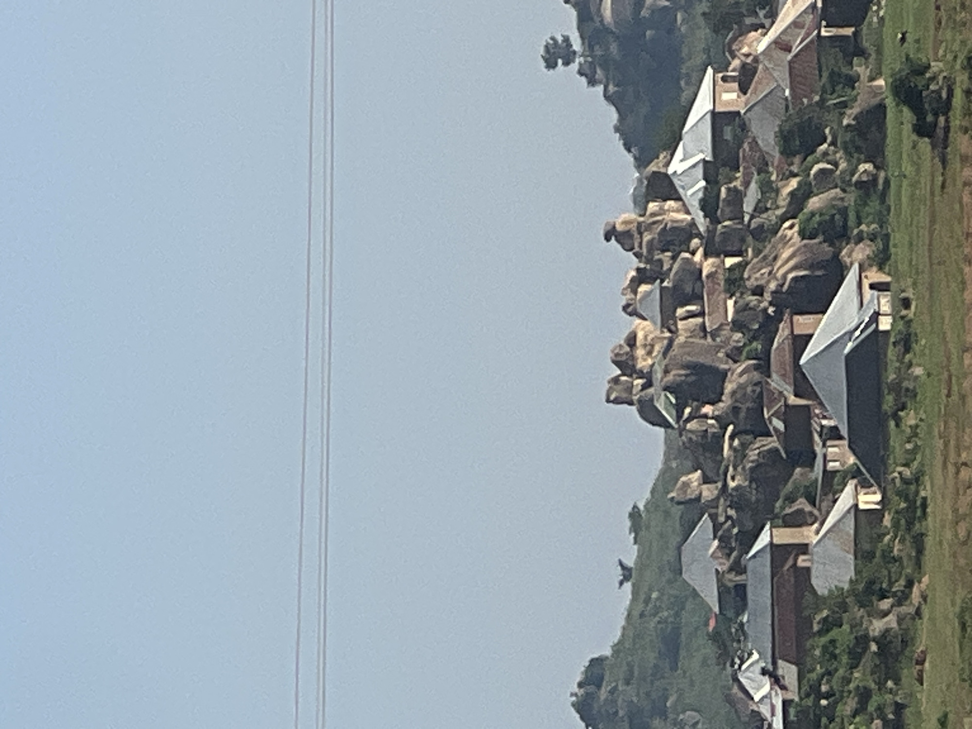 The image depicts a settlement nestled amongst a rocky, hilly landscape. Numerous buildings, characterized by their varied roof styles, primarily composed of metal and some with tiled elements, are scattered across the terrain. These structures appear to be houses, some with visible walls made of brick or plastered surfaces. Vegetation, including trees and shrubs, is interspersed throughout the scene, growing on the hillsides and around the buildings. In the distance, power lines are visible against a clear sky. The overall environment suggests a rural or semi-rural setting, possibly a village or a small town. The context provided suggests this scene is located in the city of Jos, Nigeria, a region known for its rocky outcrops and plateau landscape.