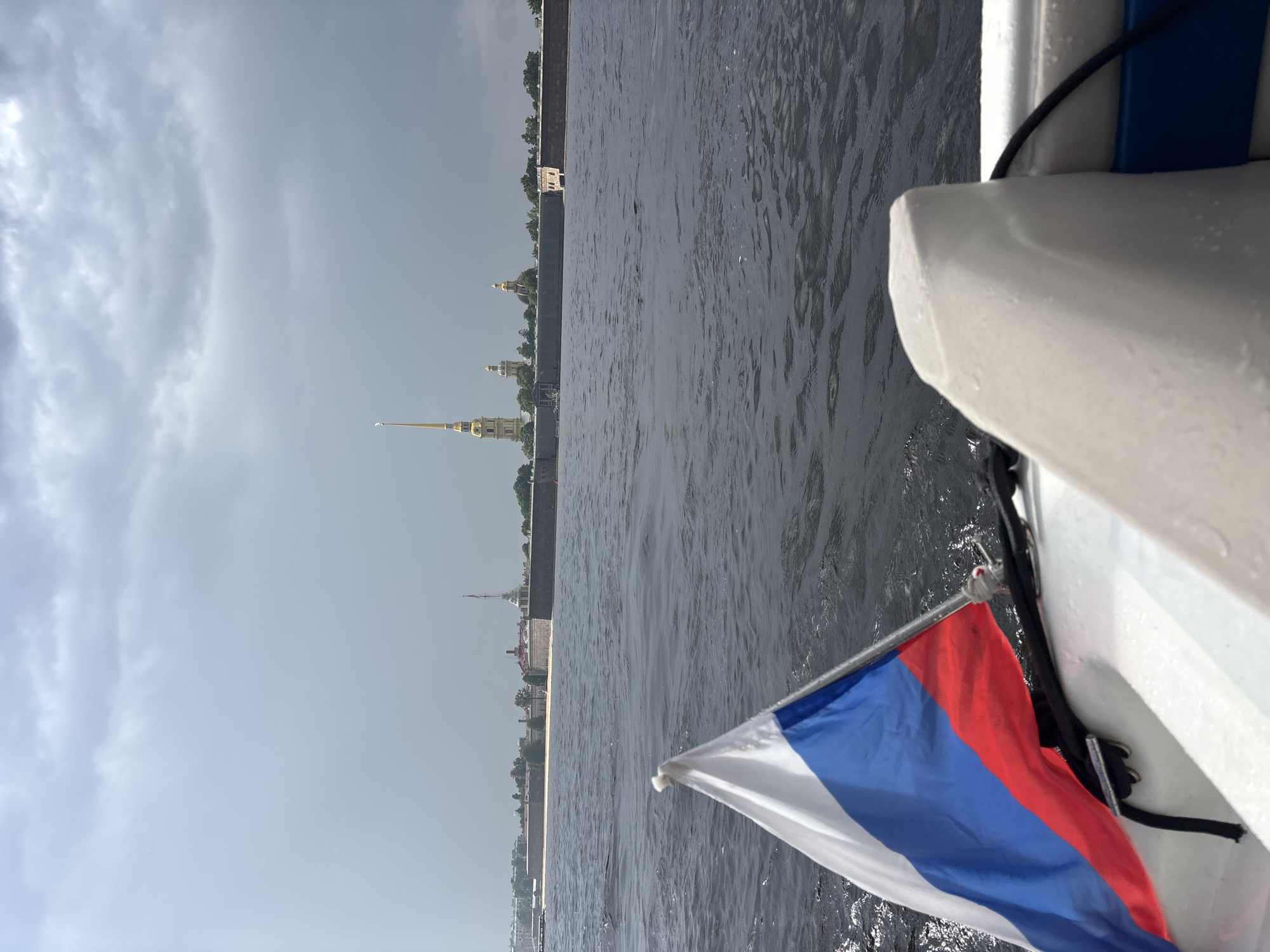 A Russian flag is in the foreground of a boat moving through dark water. In the background, the Peter and Paul Fortress in Saint Petersburg, Russia, is visible across the Neva River. The sky is overcast with grey clouds.