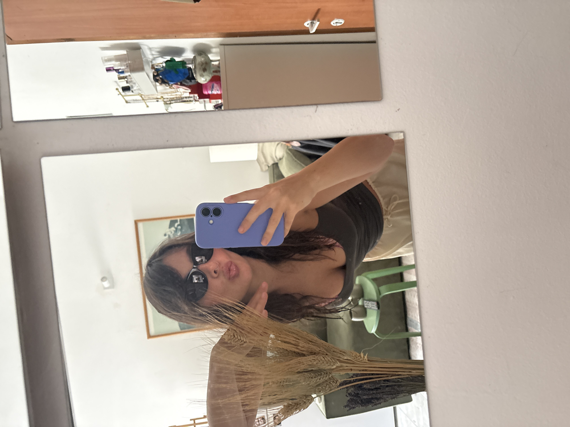 The image shows a selfie taken in a mirror. A woman with sunglasses is holding up a purple phone. Her hair is shoulder-length, and she is wearing a tank top. She is making a gesture with her hand. In the foreground, there is a dried wheat and lavender arrangement. The background reveals part of a room with a framed picture on the wall and a green chair. The location is suggested to be in Tel-Aviv, Israel, though there is no concrete evidence to confirm this from the image alone.