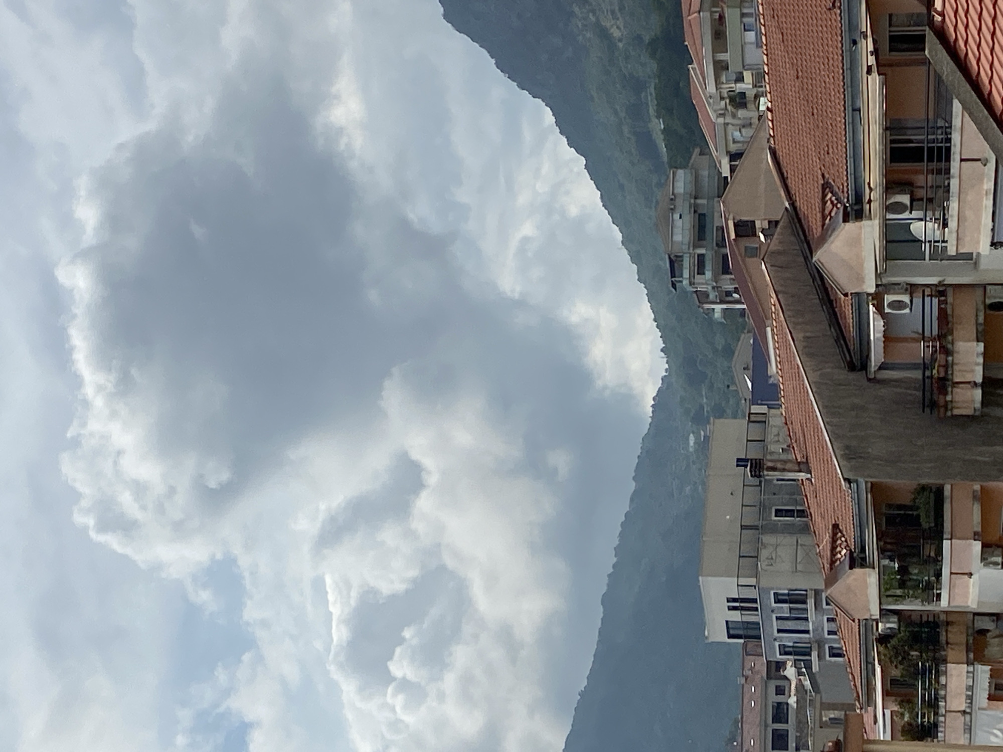 The image displays a panoramic view of a residential area situated at the base of a large mountain range, under a heavily clouded sky. The perspective suggests it was taken from a high vantage point, possibly from another building or a slope.
In the foreground and midground, a cluster of multi-story residential buildings is prominent, primarily on the right side of the frame when correctly oriented. These buildings are characterized by distinctive terracotta-tiled roofs, a common architectural feature in Mediterranean regions. Many buildings feature balconies with railings, and numerous windows are visible. External air conditioning units are mounted on the walls of several structures. The color palette of the buildings includes shades of white, beige, light brown, and grey.
Beyond the buildings, a large, verdant mountain rises steeply, forming a dramatic backdrop to the urban landscape. The mountain slopes are densely covered with green vegetation, indicating a lush natural environment. The sky is dominated by expansive, grey and white cumulus clouds, suggesting an overcast or partly cloudy day. The clouds are low, partially obscuring the higher peaks of the mountain range and casting a diffused light over the entire scene.
No people or specific activities are visible in the image, presenting a static, tranquil landscape that blends urban development with natural grandeur. The scene captures a daytime moment with diffused sunlight. There is no visible text within the image. The architectural style and natural features are consistent with the provided location context of Budva, Montenegro.