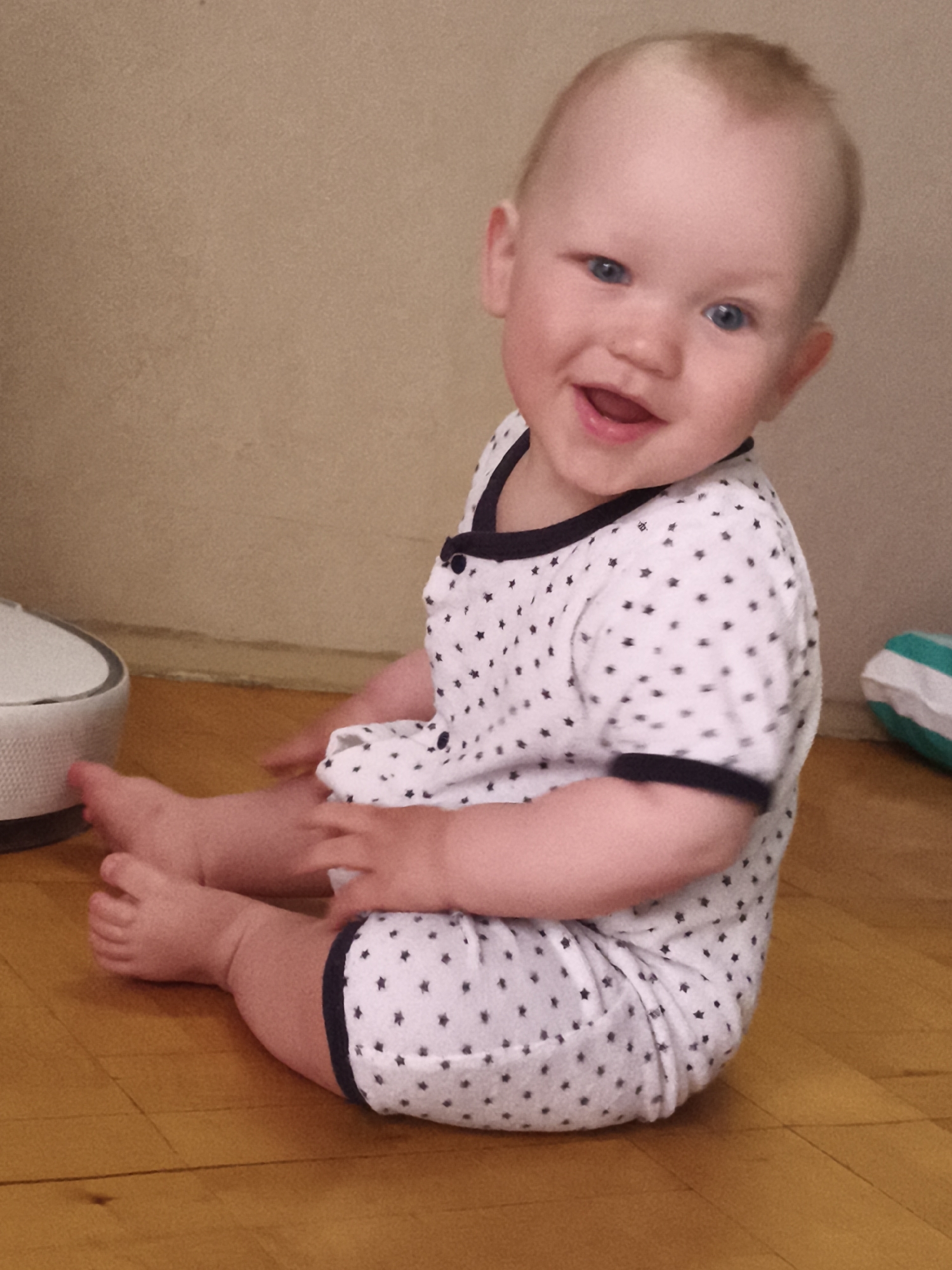 A young child with blonde hair and blue eyes is sitting on a wooden floor indoors, looking at the camera and smiling. The child is wearing a white onesie with black stars. To the left of the child is a white object with a grey rim, possibly a piece of furniture or an appliance. The background is a plain, light brown wall. There is no indication of the child's location in Chaikovsky, Russia.