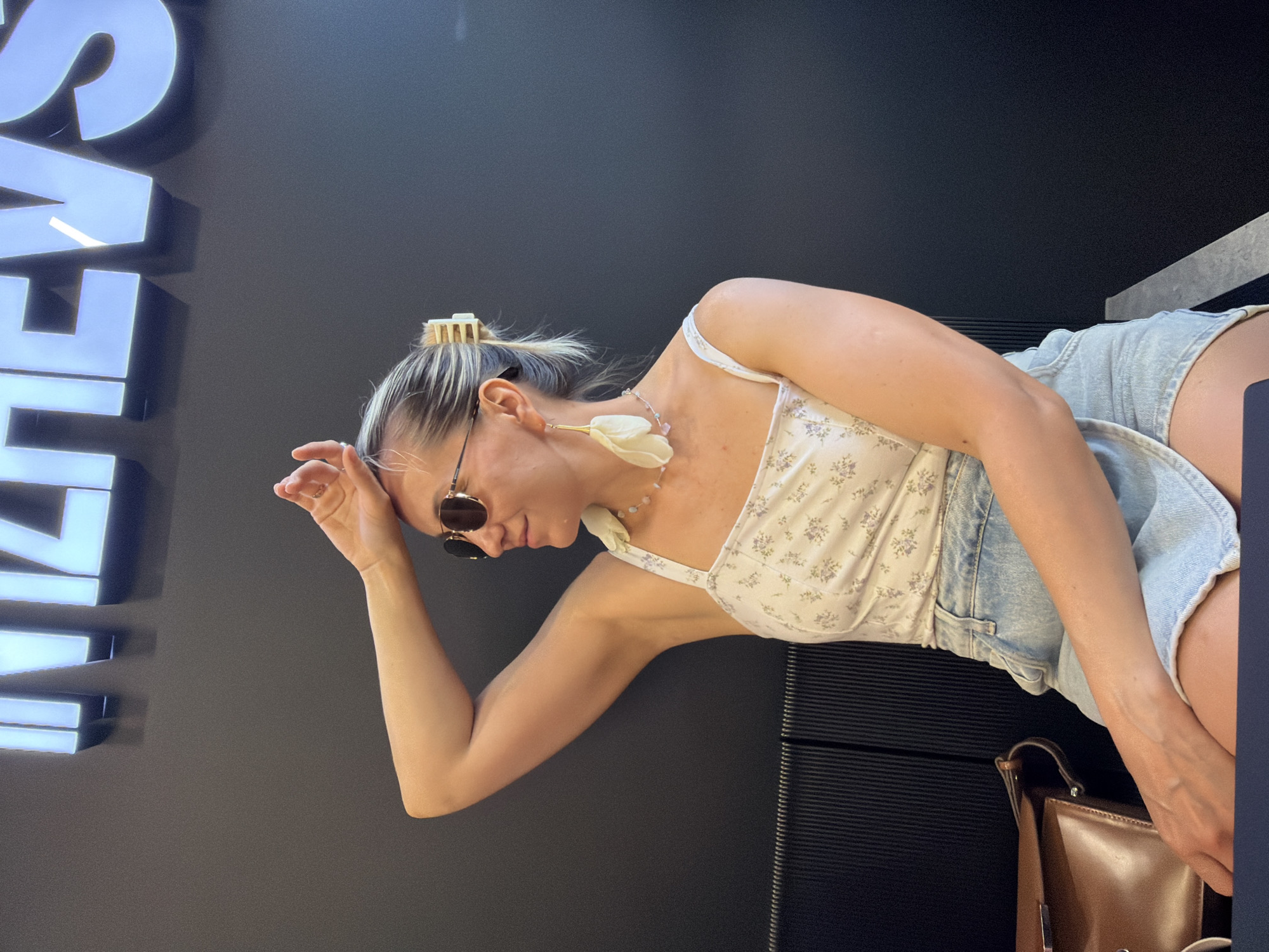 A woman with blonde hair pulled back in a bun and wearing a white floral print top and denim shorts sits with her arm raised, hand resting on her forehead. She is wearing sunglasses and large white floral earrings. A brown leather purse is visible to her left. The background is a dark wall with illuminated lettering that reads "IZHEVSK", indicating the location is the city of Izhevsk, Russia. A dark ribbed bench is partially visible behind her.
