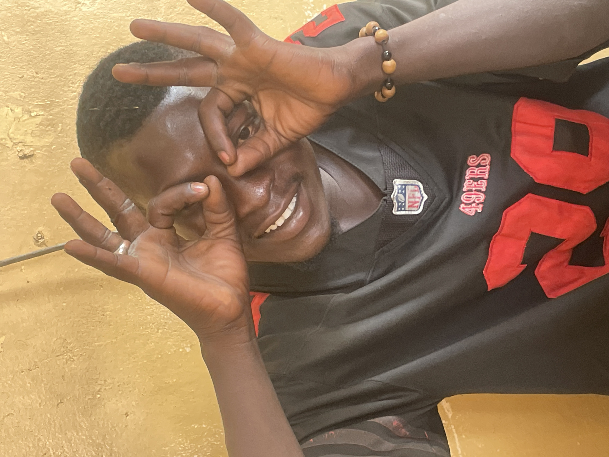 A man is indoors, wearing a black 49ers jersey with red lettering and numbers. He is making a circle shape with his fingers in front of each eye, smiling and looking at the camera. He has a bracelet made of wooden beads on his right wrist. The background is a textured, golden-yellow wall. It's not possible to verify the location in Jos, Nigeria, based on the image alone.