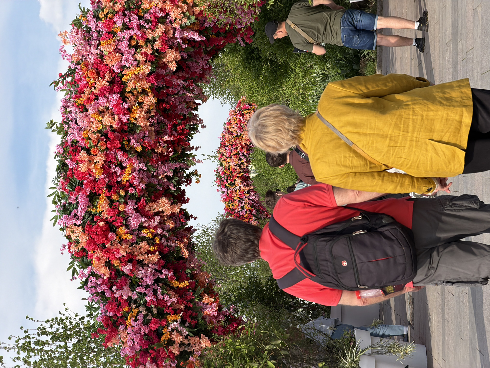 Here is a description of the image:
The image shows a group of people standing near a floral archway. The archway is densely covered in flowers of various shades of red, pink, and orange. The people are looking at the floral display. One person is wearing a yellow jacket, and another is wearing a red shirt with a black backpack. A third person is visible in an olive green shirt and jean shorts. The background includes additional greenery. It is a bright, sunny day. Based on the distinctive floral design, this image likely depicts a scene from a floral exhibition in Moscow, Russia.