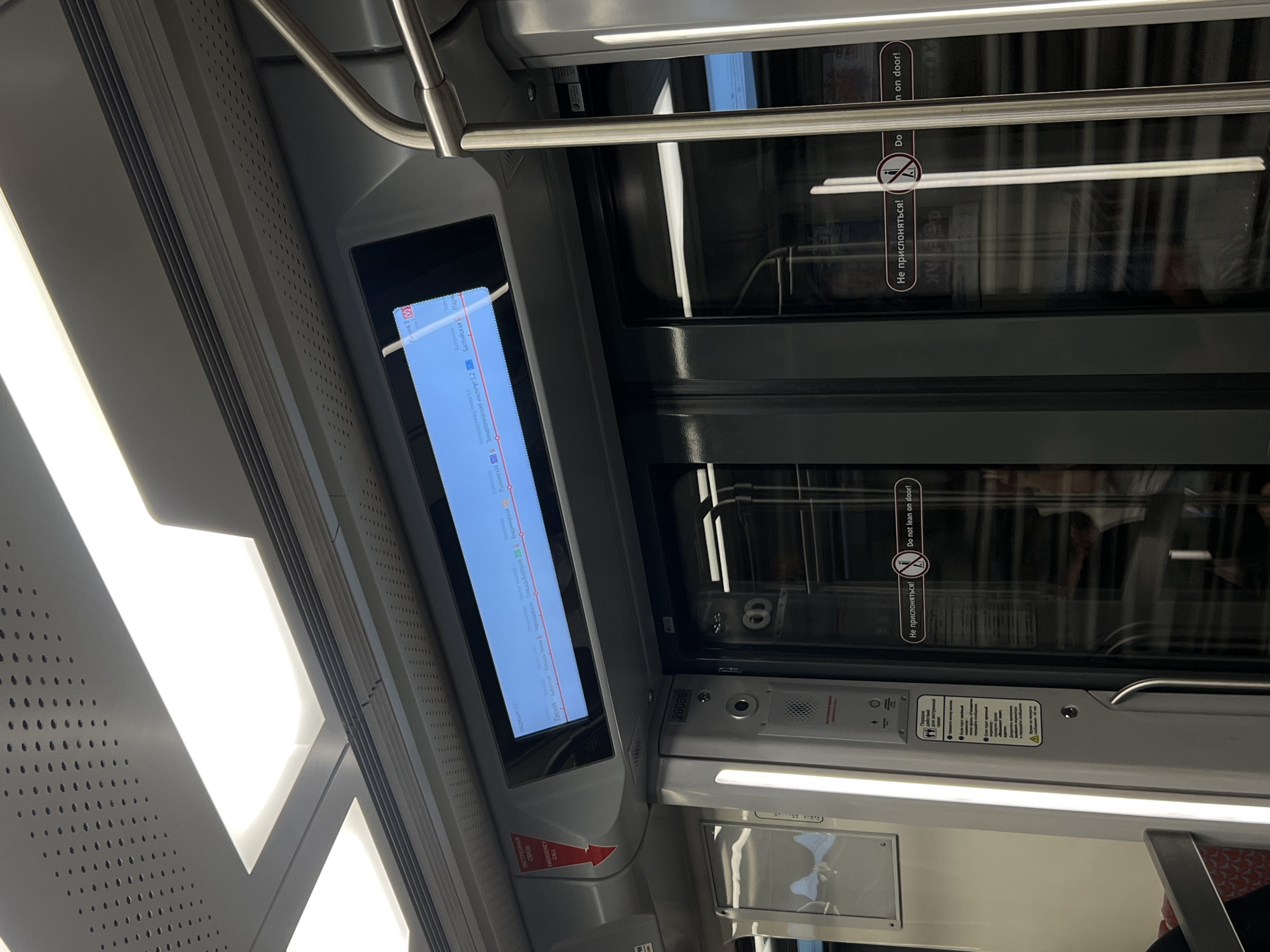 The image shows the interior of a Saint Petersburg Metro train carriage, specifically a new model with a digital route map above the doors. The route map displays a section of a red metro line, indicating "Линия 001" (Line 001) and showing a sequence of stations. The signs on the doors read "Не прислоняться!" and "Do not lean on door!". The carriage is brightly lit by overhead fluorescent lights, and the doors are reflective, showing glimpses of passengers inside. A red emergency call sign is visible on the left side of the carriage.