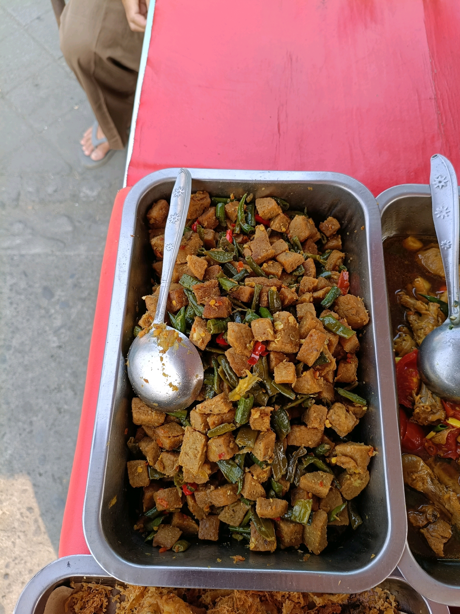At a street-side food stall in Maguwoharjo, Indonesia, a close-up overhead shot shows two metal food trays filled with various dishes. The tray on the left is predominantly filled with a stir-fry consisting of cubed tempeh, green beans, and chili peppers, seasoned with spices. A large, silver spoon rests in this tray, its bowl partially submerged in the food. To the right of this, a second tray contains a saucy dish with pieces of meat, possibly chicken, and chopped tomatoes. A ladle is visible in this second tray. The trays are placed on a red surface, likely a table or counter, which extends towards the upper right of the frame. In the background, a person's legs are visible, wearing brown trousers and grey flip-flops, standing on a grey, paved surface that suggests an outdoor setting. The lighting indicates it is daytime, with shadows cast on the ground.