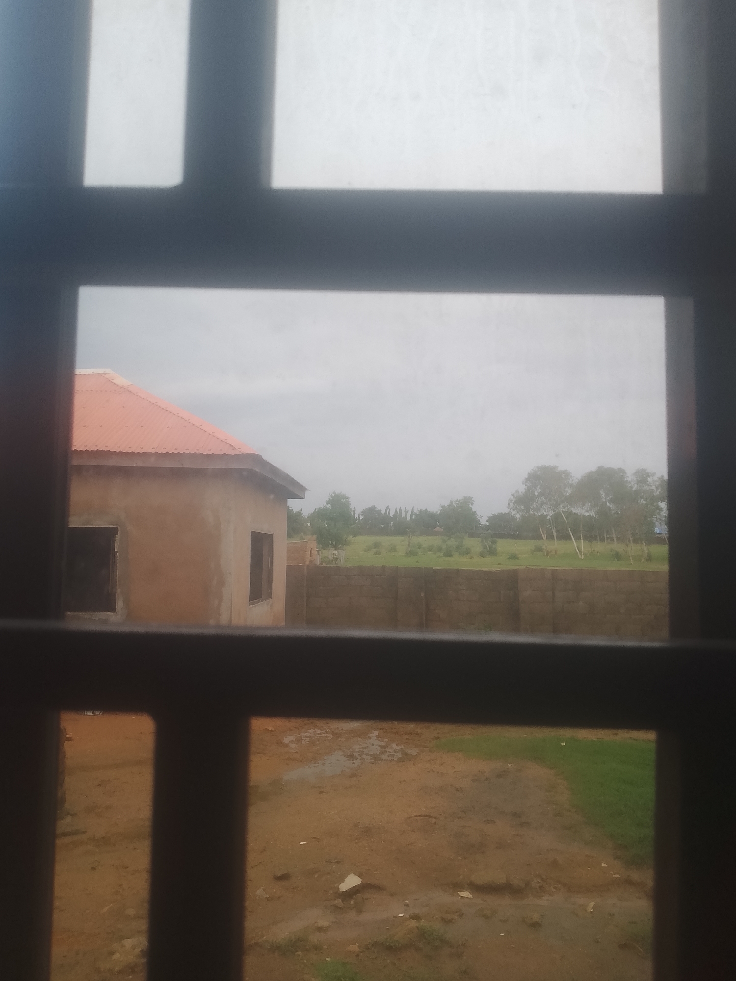 The image, captured from behind bars or a window with a grid pattern, shows an unfinished building with an orange corrugated metal roof. The building's walls appear to be made of tan-colored plaster. Two dark rectangular openings, presumably windows, are visible on the side of the building. The ground in front of the building is a mixture of mud and patchy green grass. Beyond the building, a low brick wall stretches horizontally, and further in the distance, a line of trees is visible against a cloudy sky. The scene is situated in a rural or semi-rural area. The metadata associated with the image indicates it was taken in Jauro Yinu, Nigeria.