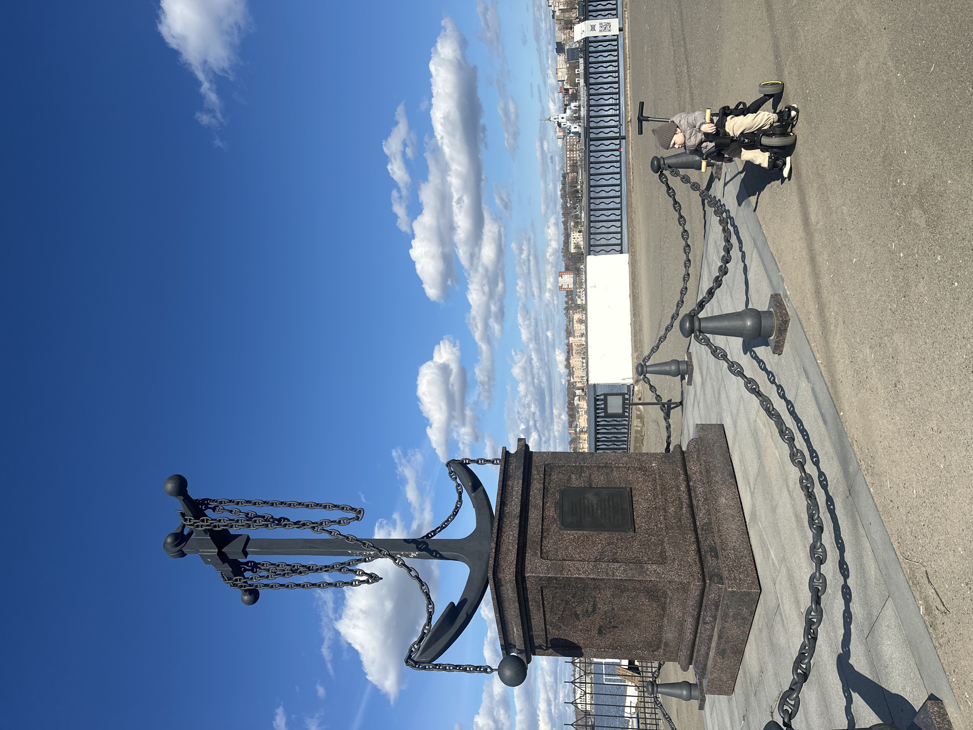 A low-angle outdoor shot shows a monument in Votkinsk, Russia, set against a bright blue sky with scattered white clouds. The monument is a granite pedestal supporting a large, dark metal anchor entwined with chains. To the right of the monument, a series of short, dark metal bollards are connected by heavy chains, forming a barrier along the edge of a concrete walkway. In the upper right corner of the image, a child in a light-colored jacket and hat sits in a stroller-like tricycle. The background reveals a cityscape with buildings and structures in the distance.