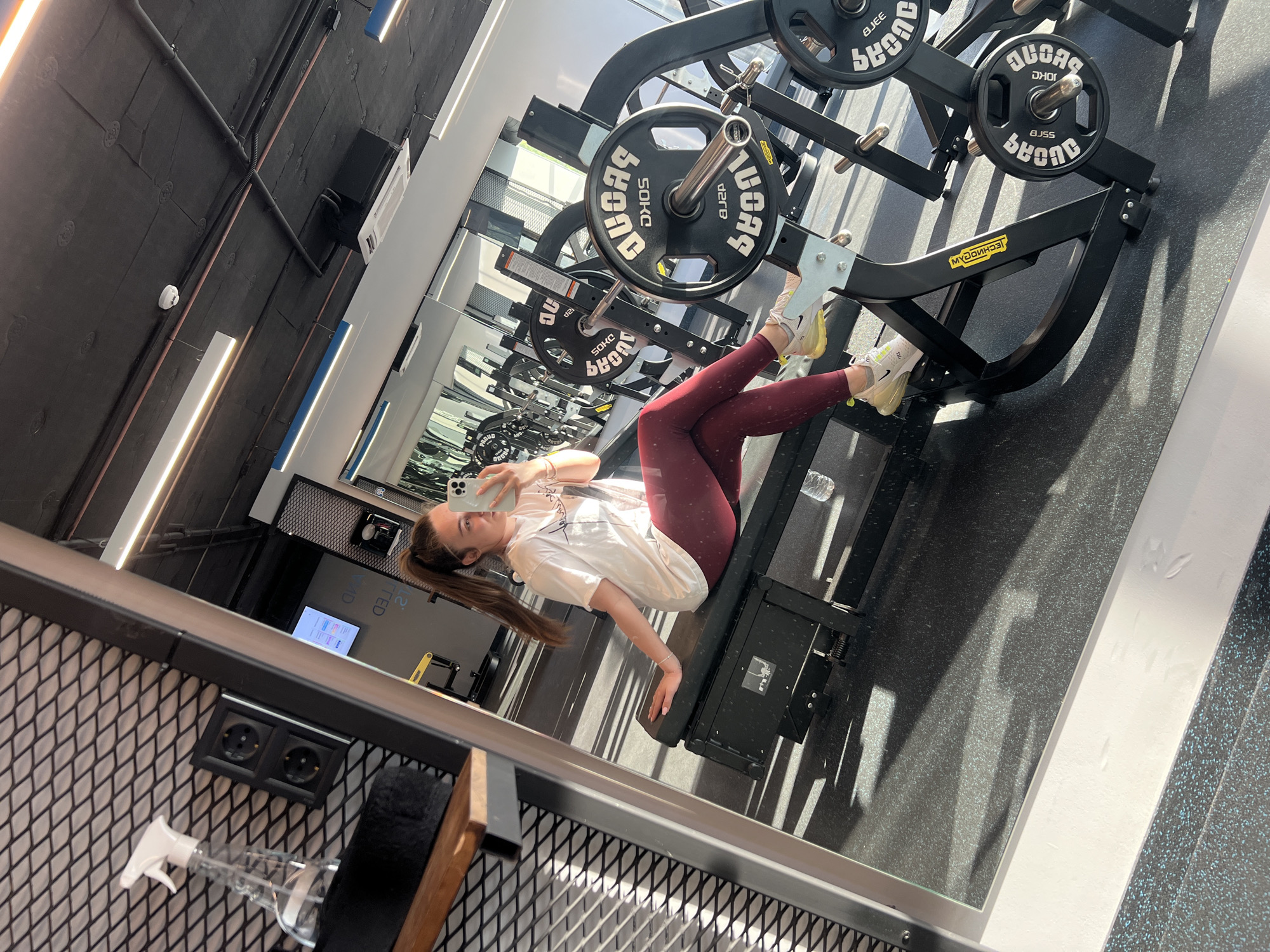 The image captures a selfie reflection of a young woman resting in a modern gym located in Plovdiv, Bulgaria. The gym exhibits an industrial design with dark, textured walls, exposed pipes, and linear light fixtures. Bright, natural light from an unseen window casts strong shadows across the gym floor, suggesting it is daytime.
The subject is a woman with long brown hair, tied in a ponytail, wearing a white t-shirt and maroon leggings. She is seated on a black leg press machine, holding a white smartphone in her right hand to take the selfie. Her expression appears neutral and calm.
The leg press machine is equipped with several black weight plates. Visible text on these plates includes "50KG", as well as various partially visible and reversed characters such as "SHONC", "LOAS", "BJSS", "DHOS", "20", "AC", and "AD", likely indicating weights or brand names like "BEYOND". The yellow logo on the machine frame clearly displays the reversed text "MYOGNHCET", which is "TECHNOGYM".
Additional objects visible in the reflection include a white spray bottle with a trigger, a black dual electrical outlet affixed to a metal mesh panel, and a clear plastic water bottle.