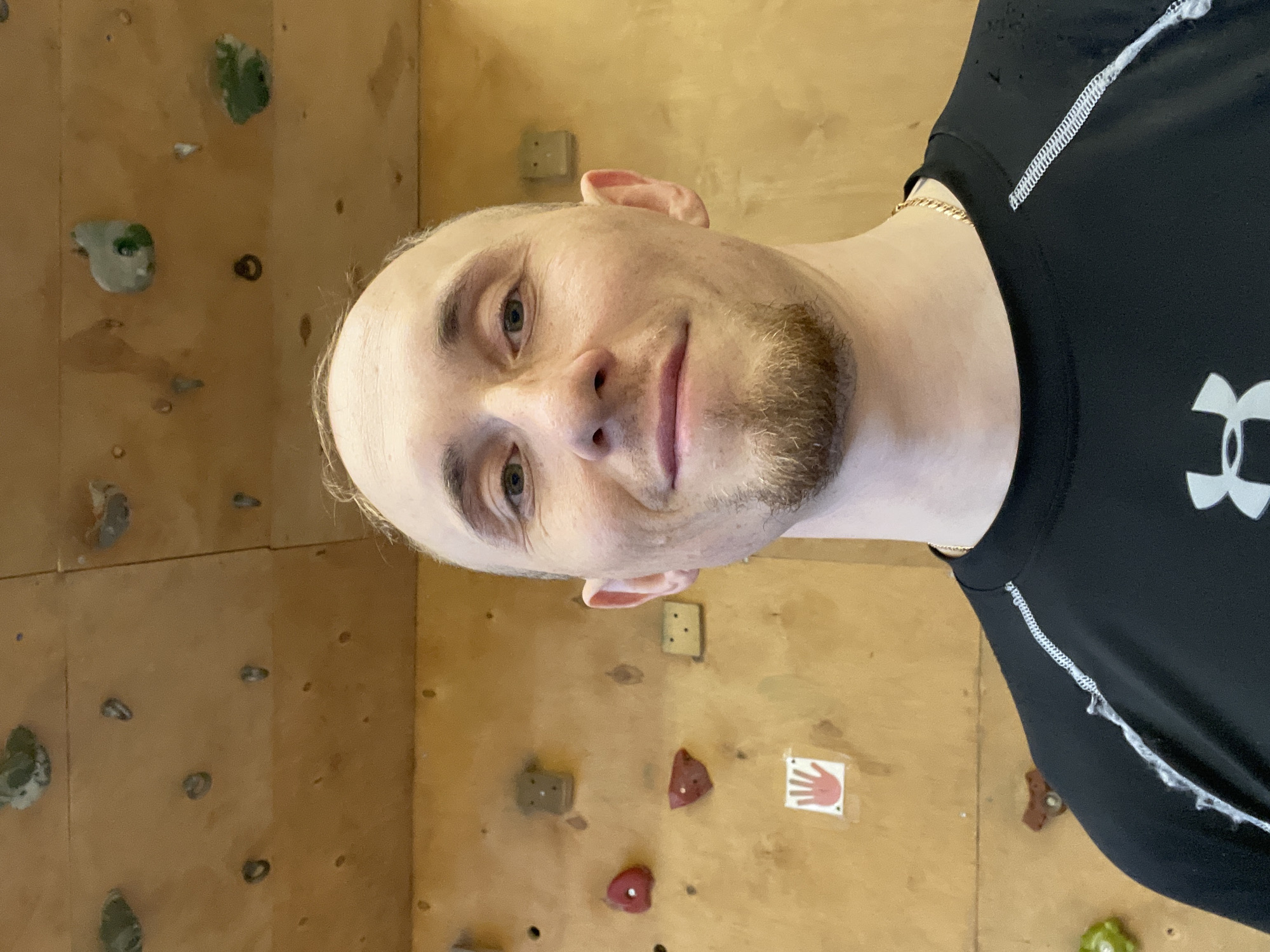 A fair-skinned man with short, light hair and a trimmed beard is positioned in front of a climbing wall. He's wearing a black athletic shirt with white trim. The wall is made of plywood and features a variety of climbing holds in different shapes and colors. Some of the holds are green, red, and beige. There is also a small sign with a red hand on it. The location appears to be an indoor climbing facility. There is no information available about the city of Sarapul, Russia.
