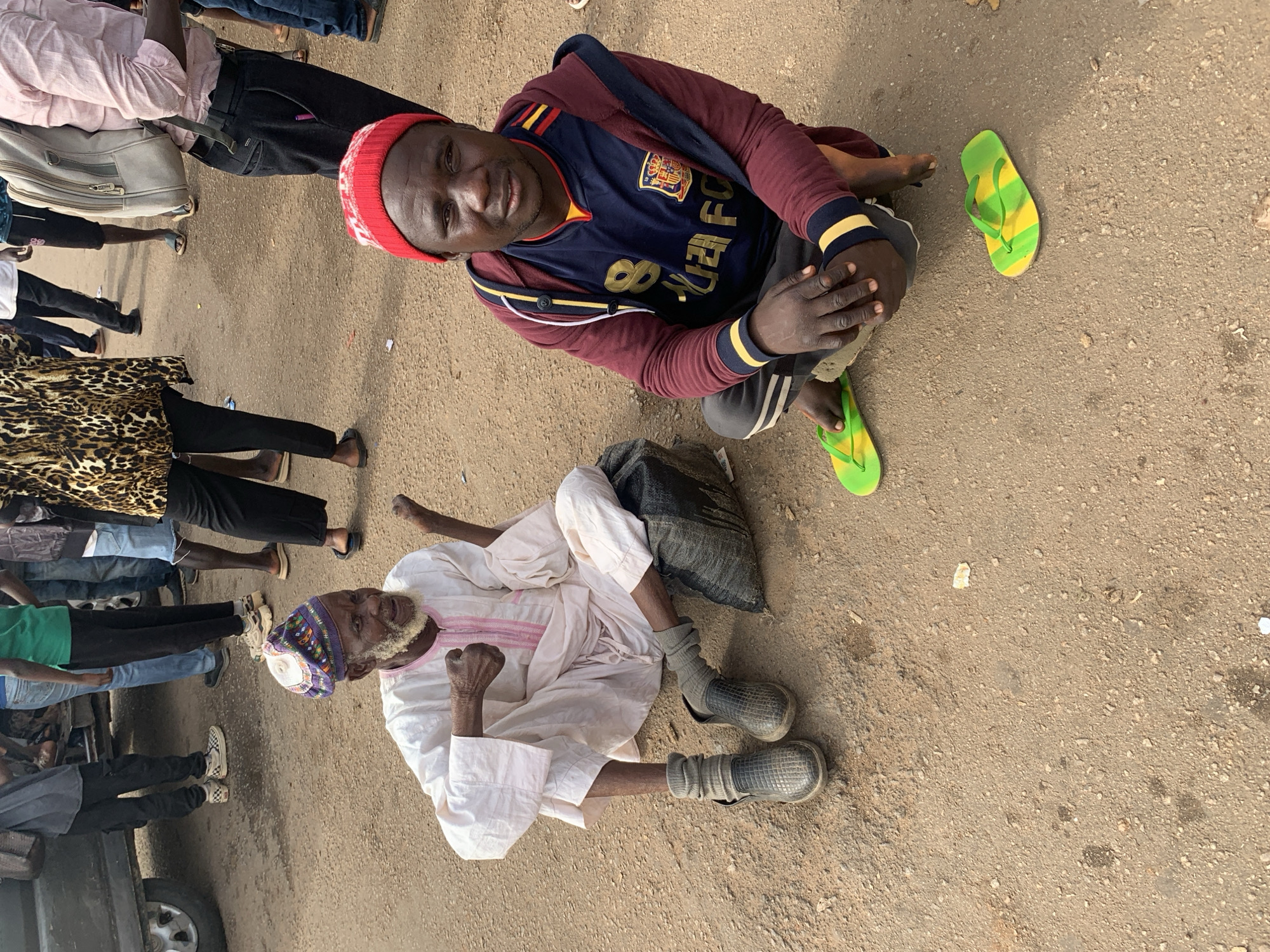 In Gura Top, Nigeria, two men are seated on the dusty ground near a vehicle. The man on the left, wearing a white robe and a patterned cap, has a white beard and appears to be speaking or gesturing. He is seated next to a dark bag. The man on the right wears a red beanie, a dark blue and red jacket, and a sports jersey. He is wearing green and yellow flip-flops. In the background, several other people are standing, some near the vehicle, and others walking away. The ground is composed of dirt and small stones, with scattered debris visible.