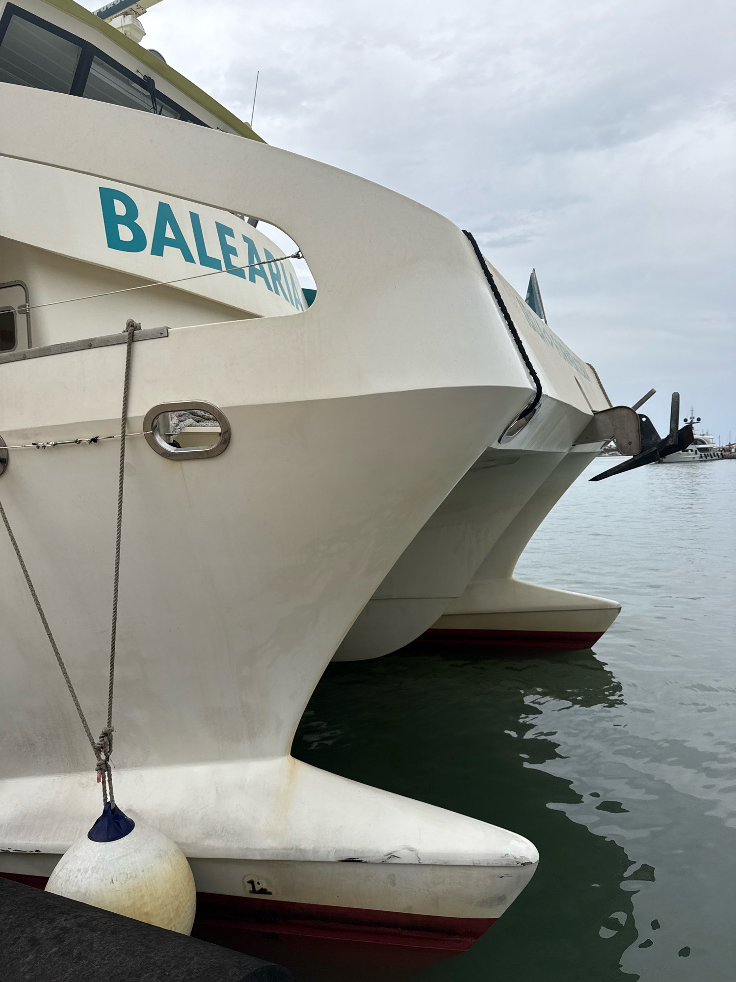 This is a medium shot, eye-level photograph of the bow of a white catamaran ferry, identified by the name "BALEARIA" emblazoned in teal lettering on its hull. The ferry is docked in the harbor of Ibiza, Spain. The catamaran's two hulls are visible, separated by a narrow channel of dark, rippling water. A thick, gray rope extends from the ferry to a large, white cylindrical fender floating on the water, which is partially obscured by a black rubber fender or dock edge in the foreground. An anchor mechanism is visible attached to the bow, with a black anchor pointing downwards. The sky is overcast with light gray clouds. In the background, out of focus, are other vessels and structures, suggesting a busy port environment. The overall impression is of a modern ferry at rest in a Mediterranean port.