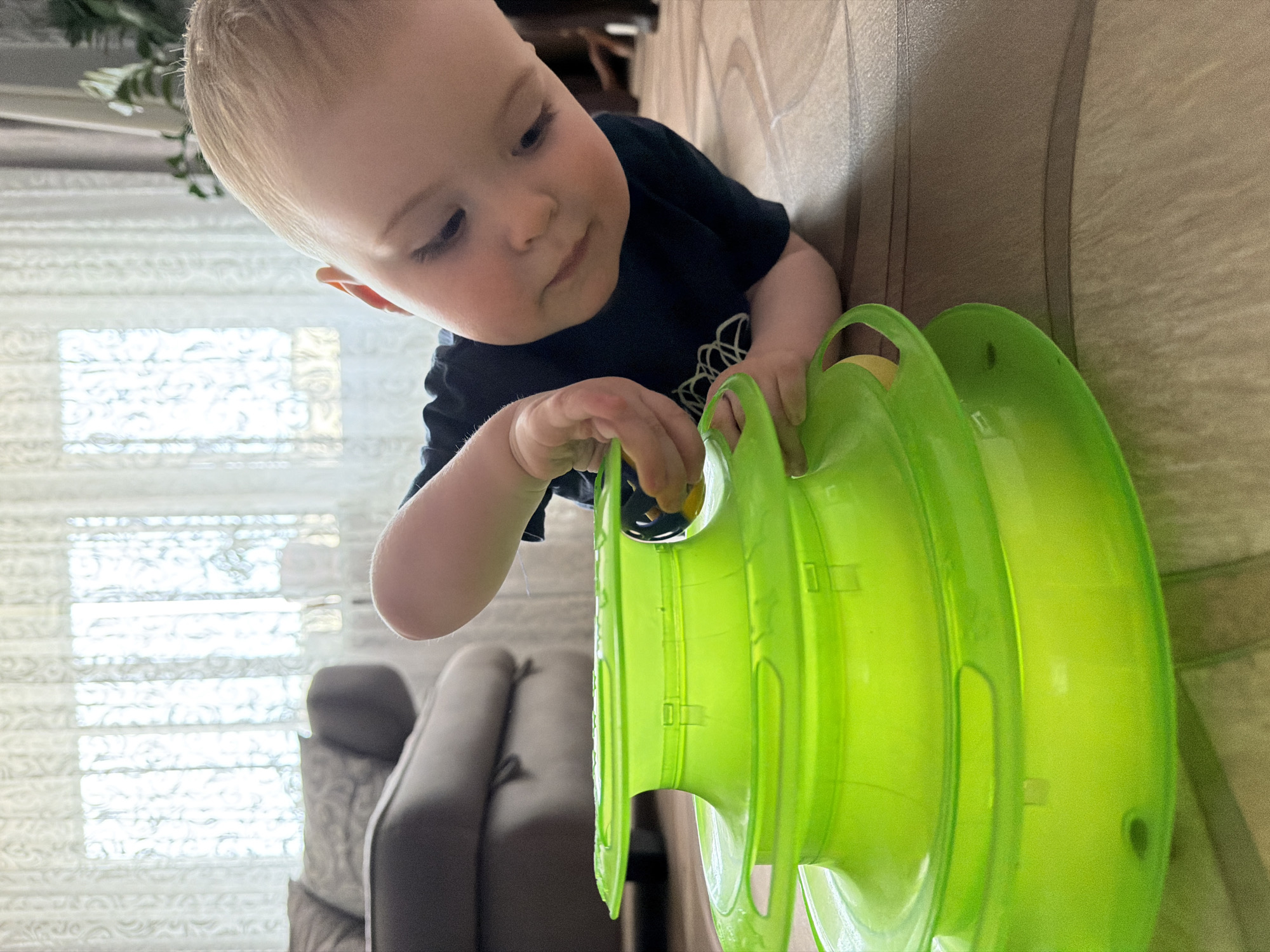 A toddler is playing with a three-tiered, green cat toy on a table. The child is wearing a dark-colored shirt and has light hair. The table has a wood-grain pattern. In the background, there is a window with blinds and a gray couch. The location cannot be determined from the image and does not appear to be in Votkinsk, Russia.