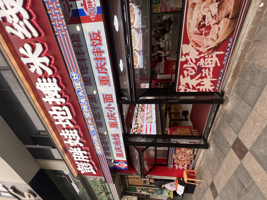 The image displays the storefront of "Chubby Baby Street Stall Rice Noodles" (憨胖娃地摊米线), a restaurant located in Jiangjin, Chongqing, China. The establishment features a prominent red facade with large, illuminated white and red Chinese characters. Below the main sign, additional white signage advertises "Chongqing Rice Noodles," "Chongqing Small Noodles," and "Chongqing Mixed Rice." Smaller text on the main sign mentions "Deep Clay Pot Three Delicacies" and "Insist on Fresh Stir-fry."
The restaurant entrance consists of large glass panels, with the left section hinged upwards, forming an open window. Inside, the brightly lit interior reveals a dining area furnished with multiple wooden tables and stools. Large digital menu screens display various food items with images and prices. Towards the rear, a serving counter is visible, where at least two staff members, one wearing a blue apron, are engaged in food preparation or service.
On the tiled sidewalk outside, a male individual wearing a dark jacket stands to the left of the entrance, holding a white plastic bag and looking upwards. Adjacent to him is a black crate topped with oranges and a small wooden stool. The sidewalk is paved with grey and brown checkered tiles. A large red-and-white promotional banner, depicting a hotpot-style dish with assorted meats and vegetables, is affixed to the lower part of the right-hand window. The building to the immediate left of the restaurant has a grey facade, featuring a white sign with green text displaying the phone number 37983776606. The sky appears overcast.