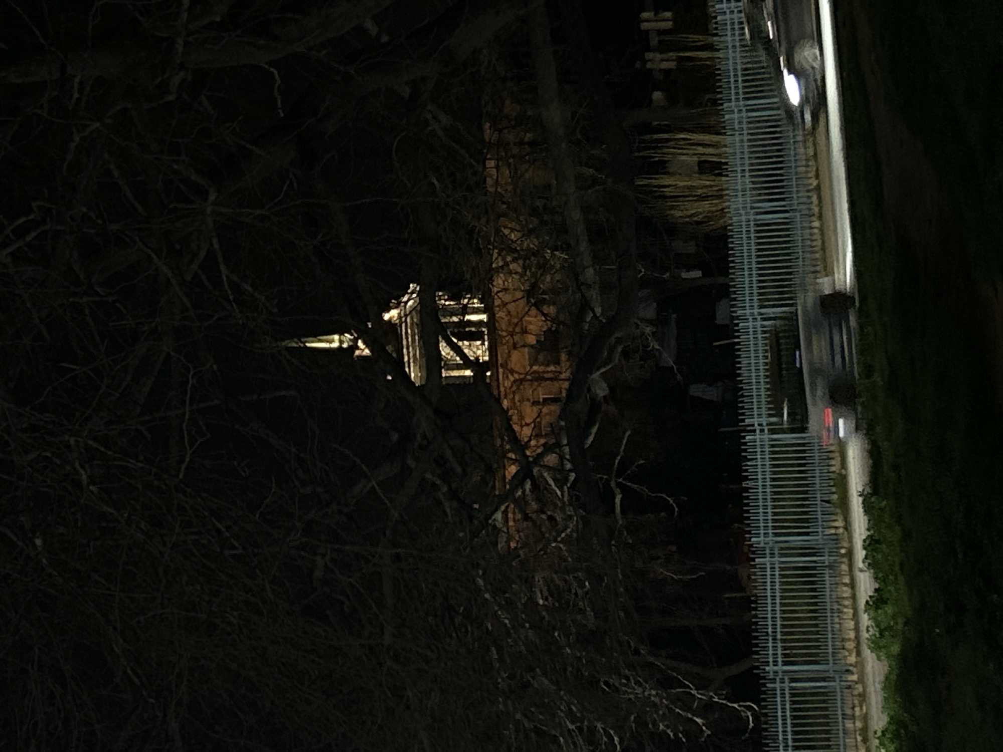 The image, taken at night, shows a building framed by bare tree branches on the left side of the frame. The building is brick and has a steeple. To the right is a metal fence and a road. The location is Varna, Bulgaria.