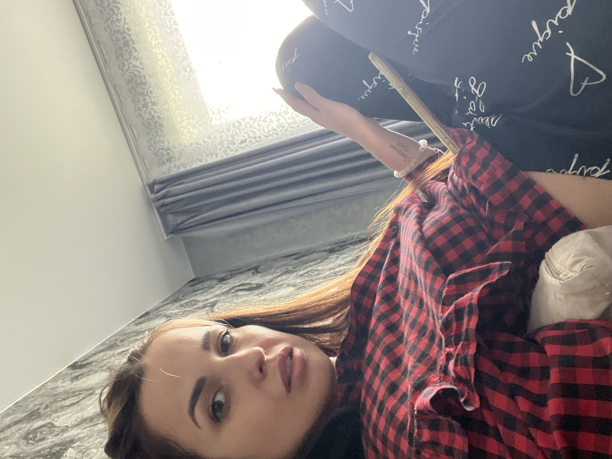 A low-angle shot shows a woman lying down, possibly on a couch or bed. She is wearing a red and black checkered shirt. Her left arm is raised, holding what appears to be a tablet or phone. The background includes a window with curtains and a wall with a patterned surface. The image appears to be taken indoors. The location details are unknown.