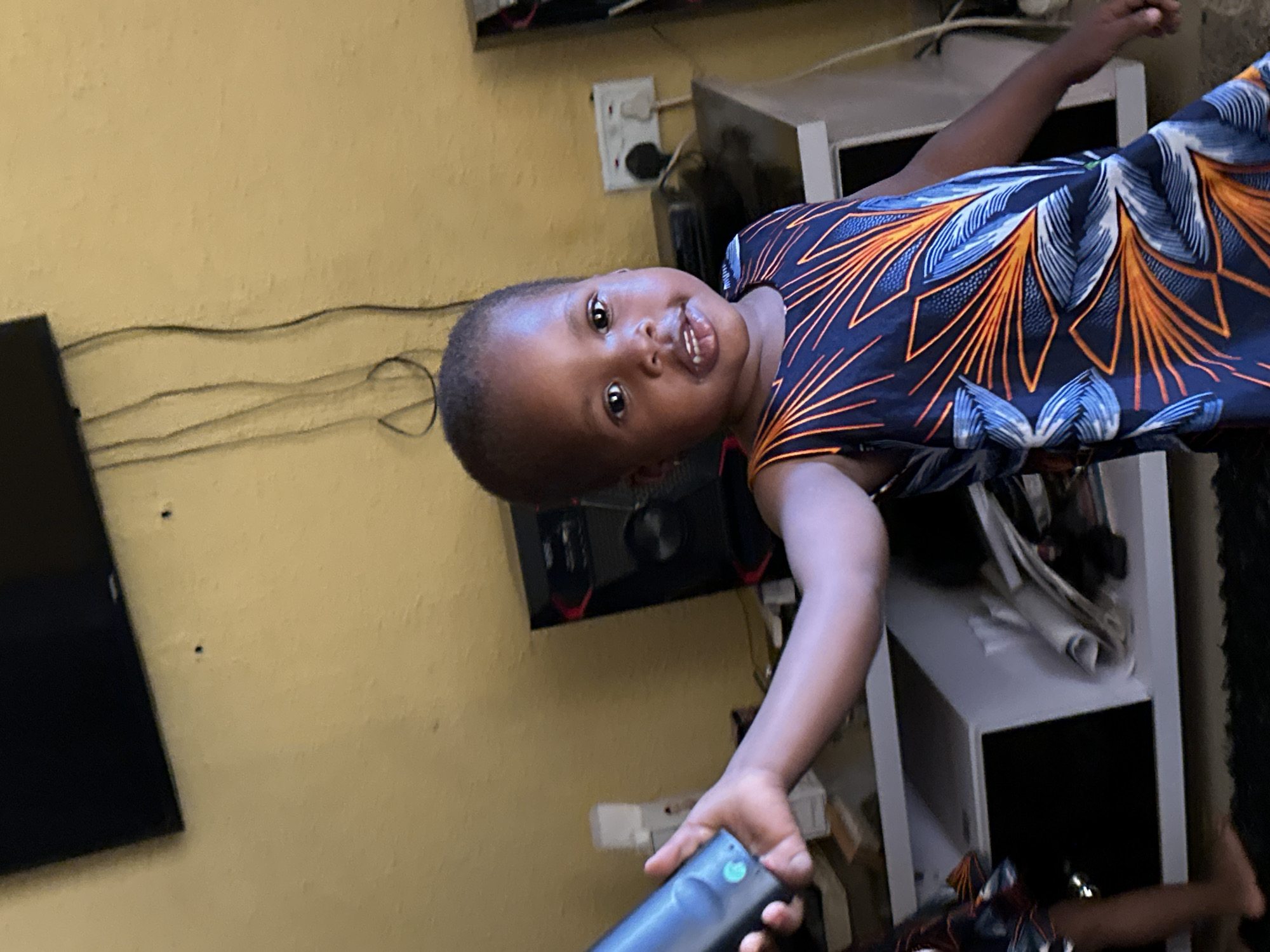 The image shows a young child wearing a dark blue dress with an orange and blue patterned design. The child is looking towards the camera with a slight smile, showing their teeth. The child's head is tilted to the side. The child appears to be in a room with yellow walls. There are wires visible on the wall behind the child, as well as what appears to be an electrical outlet. In the background, there is a black television screen to the left and a white shelving unit to the right. A hand holding a black remote control with a green button is visible in the lower right portion of the image. The image is taken from a low angle. The overall impression is of a candid, indoor photograph.