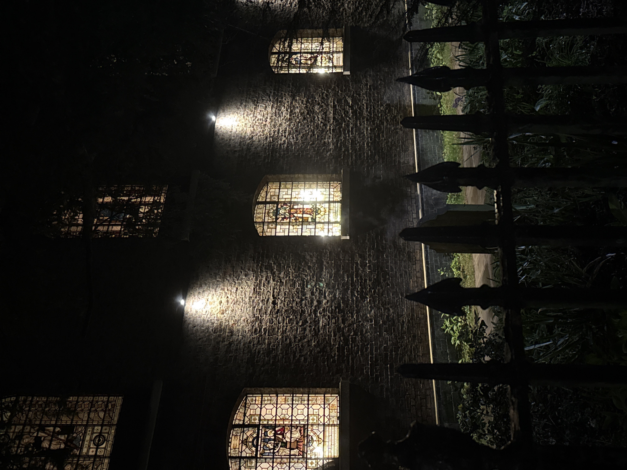 The image displays the exterior of St. Giles Cripplegate church, situated in the City of London, United Kingdom, during nighttime. A dark, textured brick wall forms the main subject, featuring five visible arched stained-glass windows across two levels. Each window is internally illuminated, revealing religious scenes and geometric patterns in various hues. Three individual upward-facing spotlights are affixed to the brickwork, casting focused light beams onto the wall directly above three of the lower-level windows. A black, wrought-iron fence with pointed finials extends horizontally across the foreground, partially obscuring a dark garden area containing green foliage and obscured ground cover between the fence and the church wall. No individuals are present, and no actions are observed.