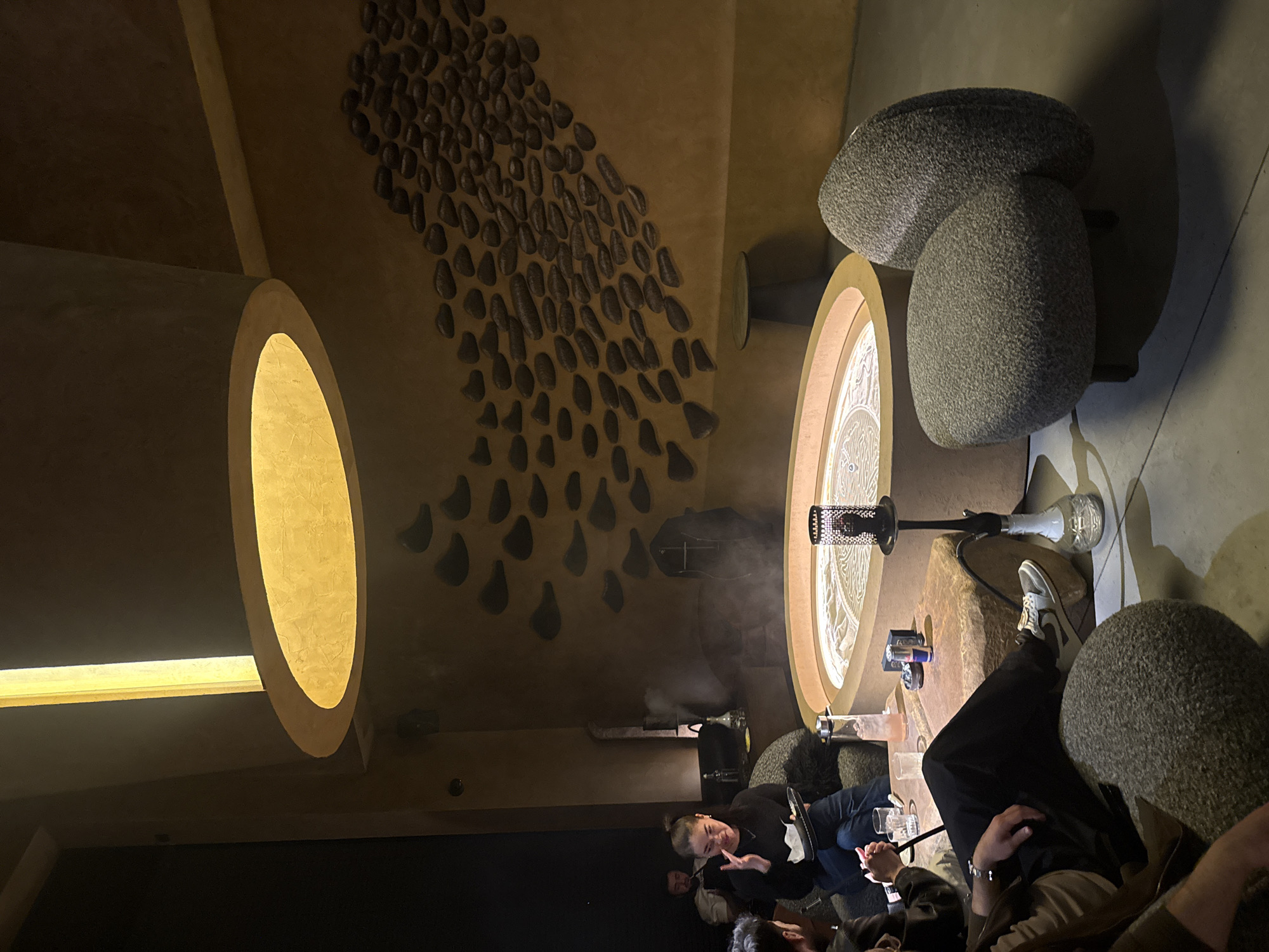 The image shows an indoor scene in what appears to be a hookah lounge. The setting includes unique decorative elements: textured wall decorations resembling stones or shells, illuminated oval fixtures, and pebble-like seating. A hookah is centrally placed, with smoke emanating from it. Several people are present, sitting around the hookah and engaged in conversation. The environment is dimly lit, contributing to the lounge atmosphere.
While the image location is specified as Khimki, Russia, there are no forensic details in the image to confirm this location. The description focuses on observable elements within the frame.